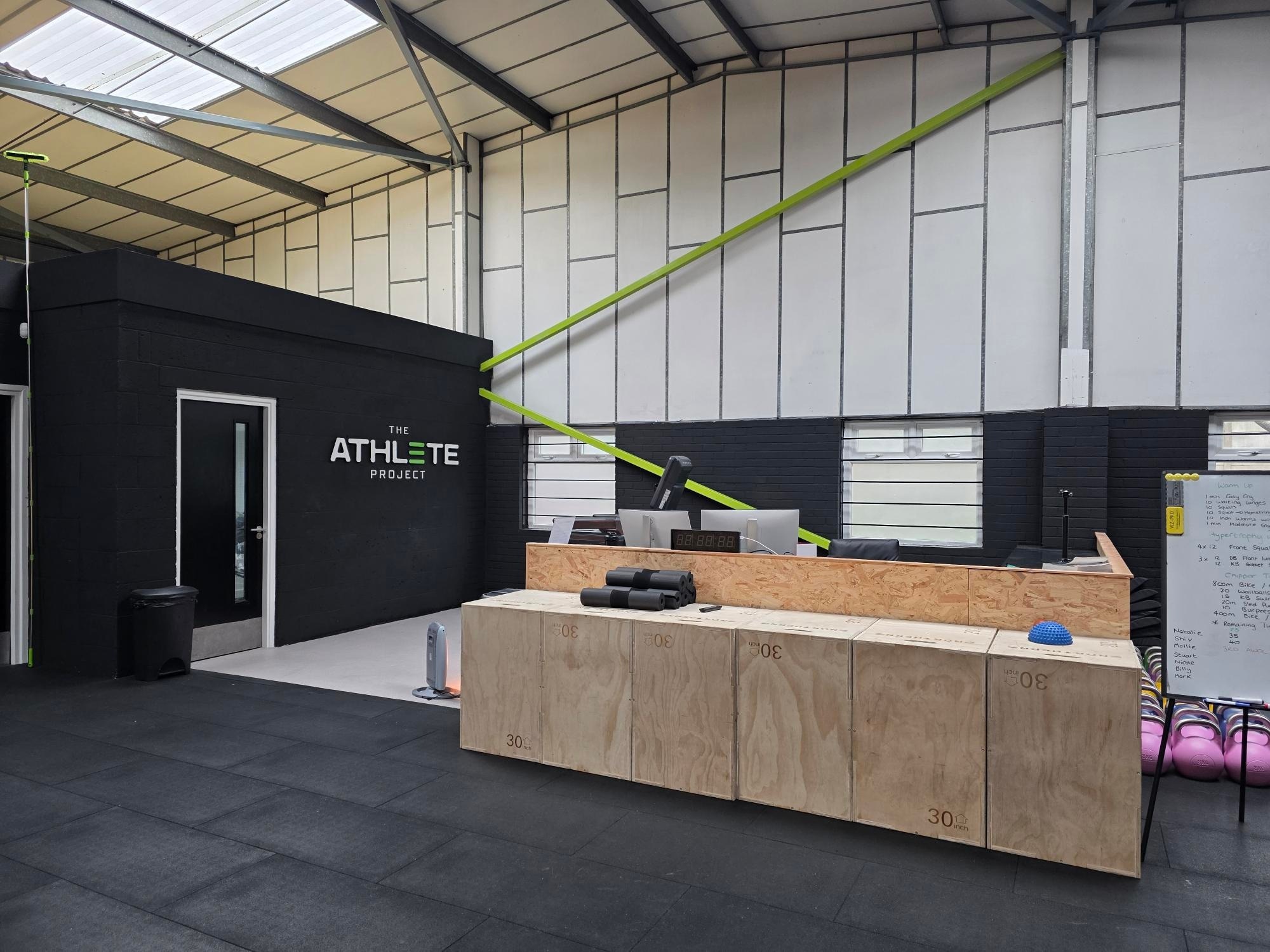 Interior of a fitness facility named "The Athlete Project" featuring a reception area with a wooden counter and exercise equipment.
