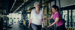 Two women taking part in a Exercise referral course at a gym. One woman is older and is exercising with dumbells while the younger women assists her.