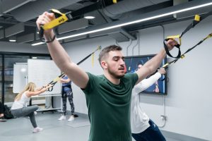 A male student learning on the suspension fitness TRX course in a fitness classroom during his CPD module. A tutor is standing in the background observing