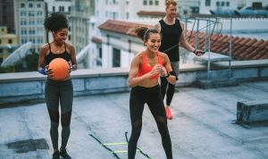 Smiling fitness women and a man doing fitness training on terrace. Man doing workout using skipping rope while his female colleagues training with agility ladder and basketball.