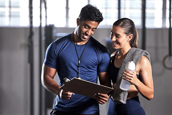 A fitness coach in a blue shirt holds a clipboard while discussing training with a female athlete, who holds a water bottle and towel.