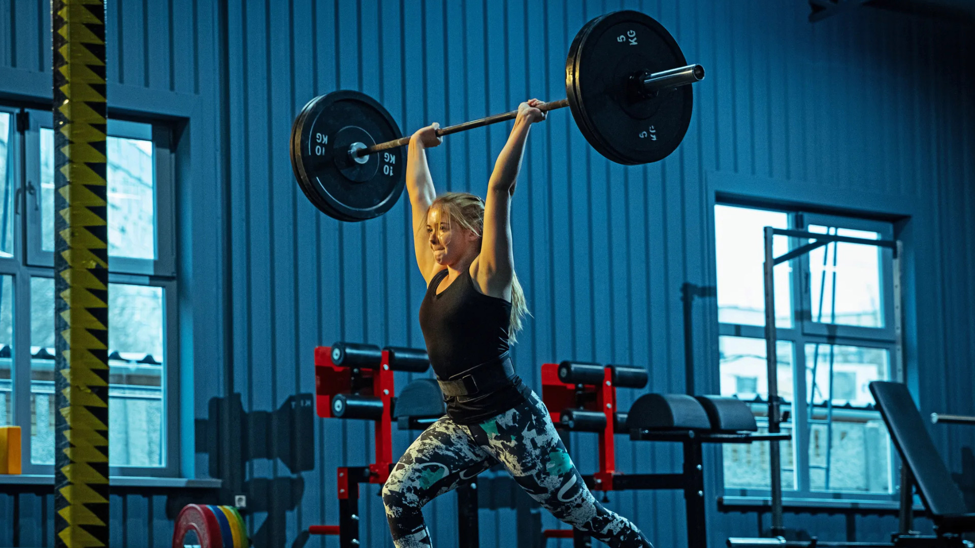 A person performs a barbell overhead lift in a gym, showcasing strength amidst a backdrop of fitness equipment and blue walls.