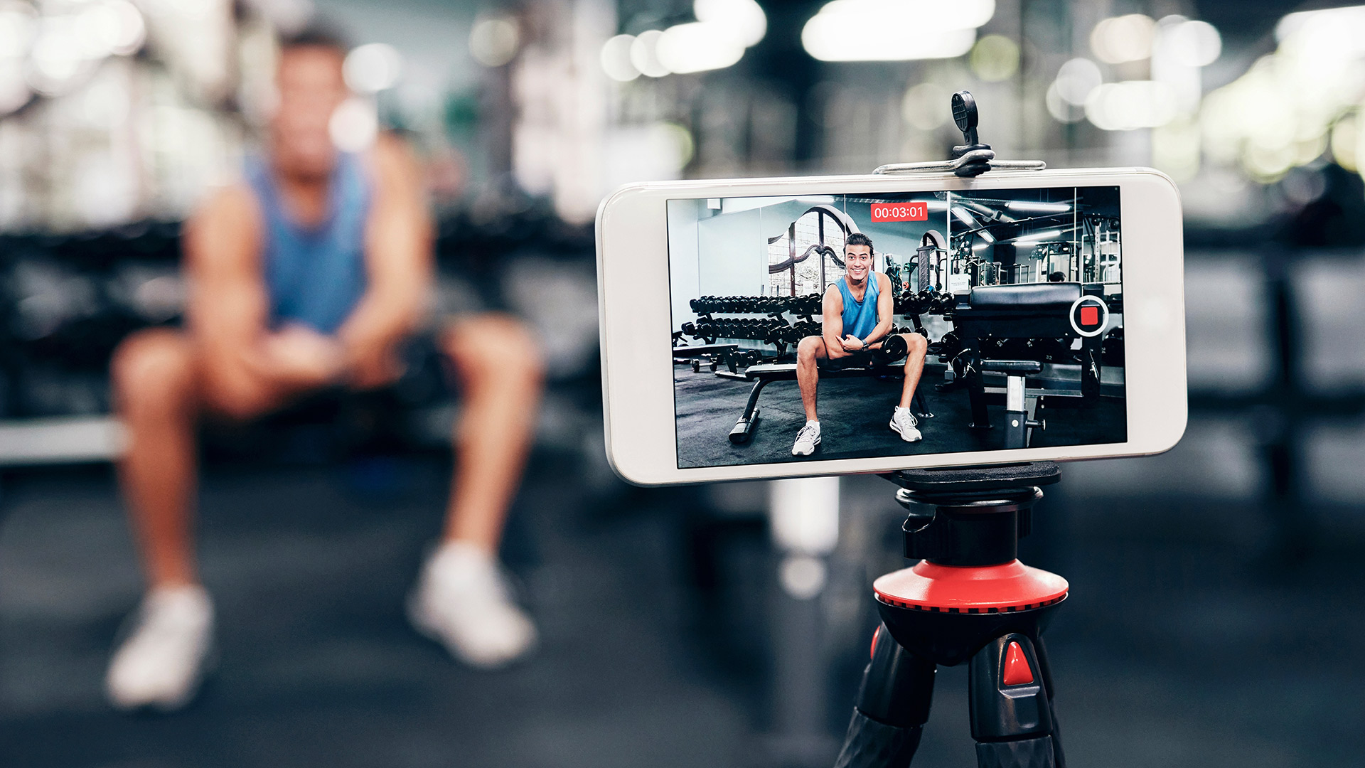 A smartphone on a tripod records a person exercising in a gym surrounded by weights and fitness equipment.