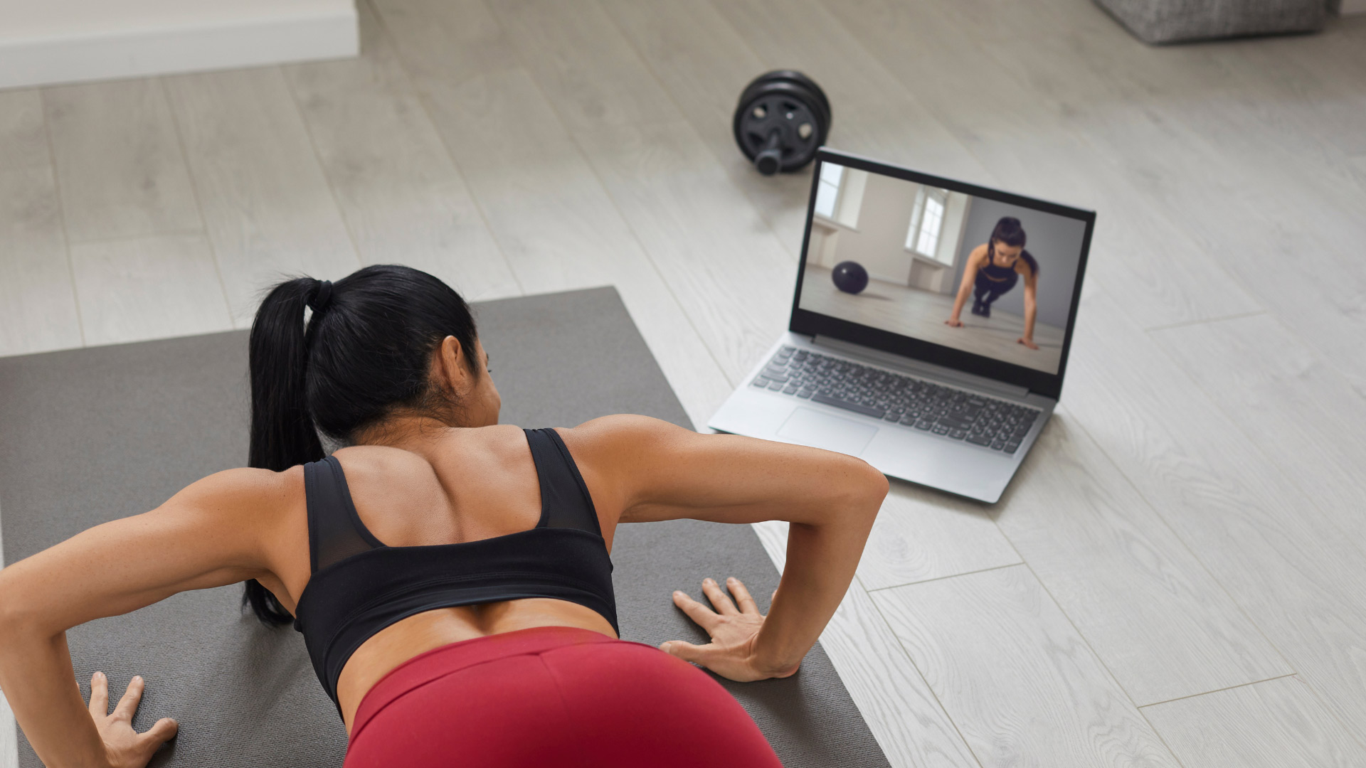 A woman performs push-ups on a yoga mat at home, following an online workout displayed on a laptop. A dumbbell rests nearby.