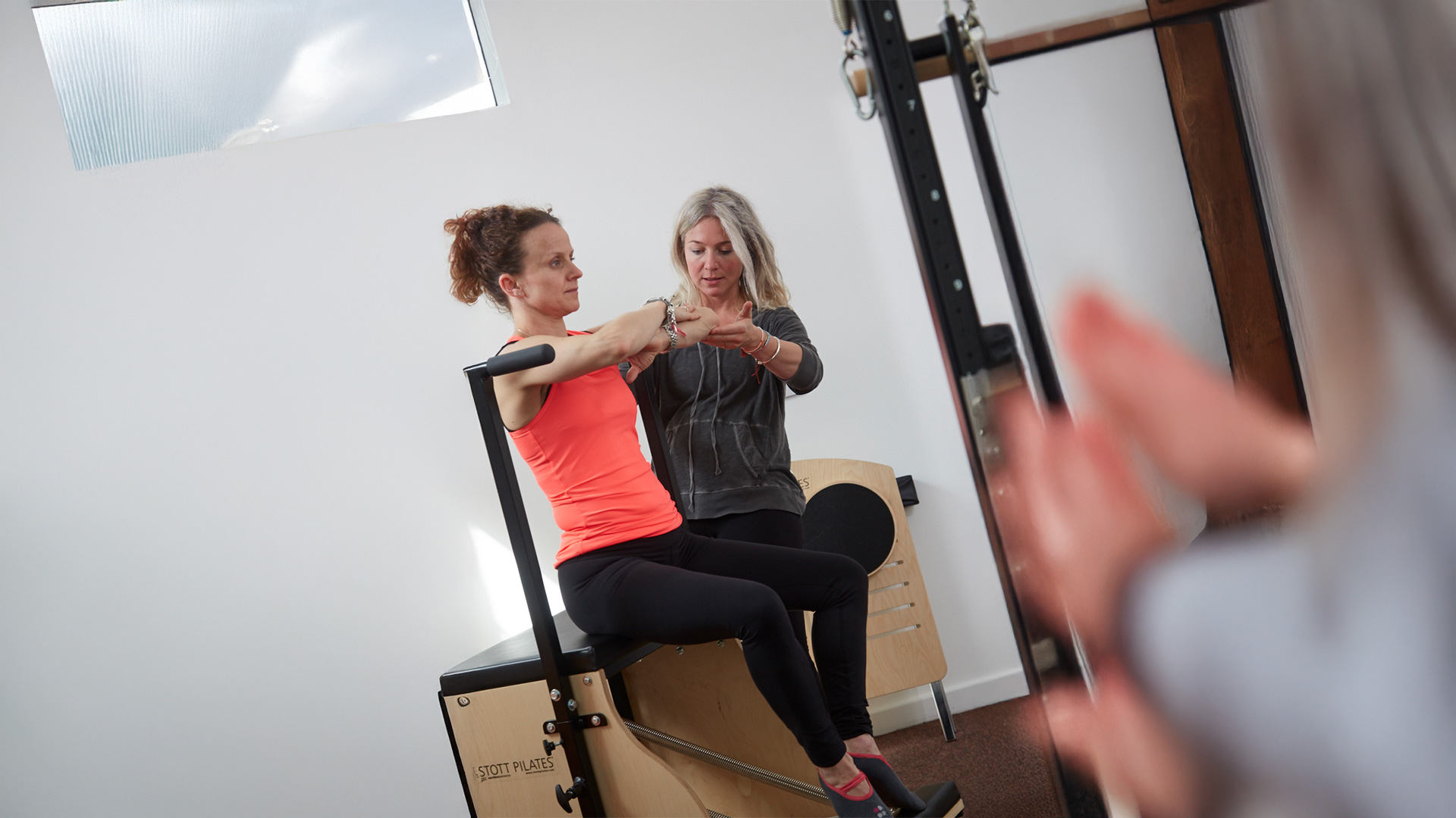 A woman in a red tank top practices Pilates on a reformer, guided by an instructor in a studio with a mirror and wooden equipment.