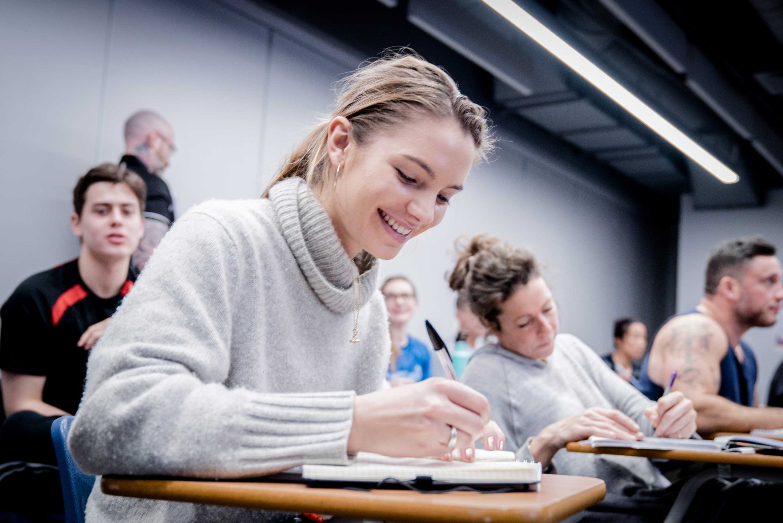 A student writes in a notebook during a class, surrounded by peers seated at desks in a modern classroom setting.