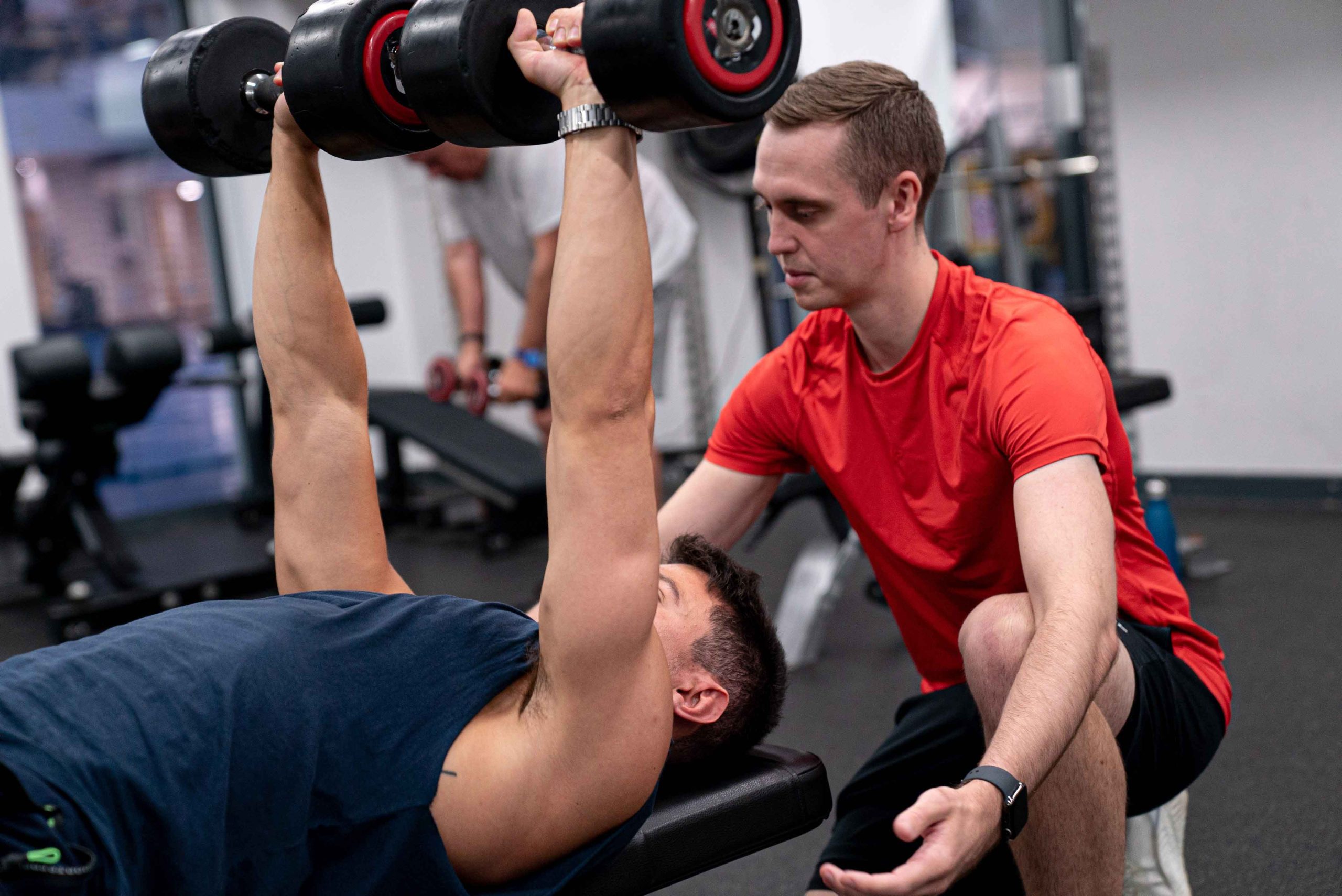 A person lifts a dumbbell on a bench while another offers guidance in a gym setting. Weight equipment and a bright environment are visible.
