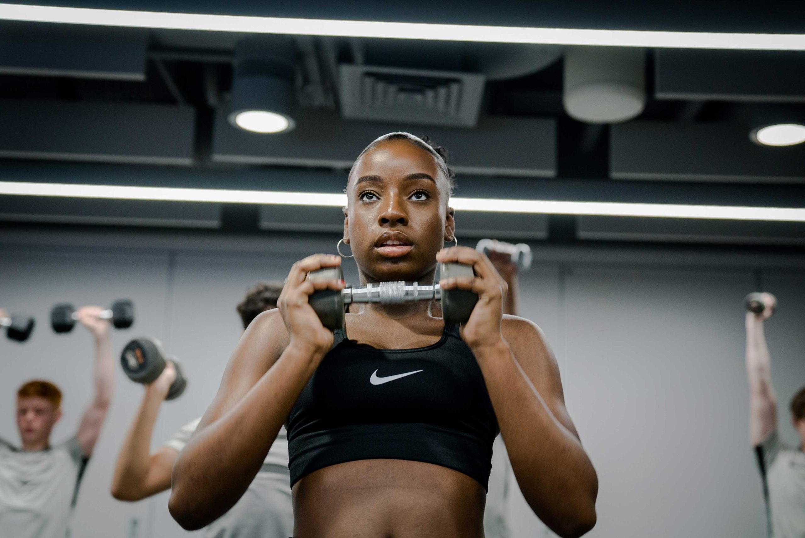 A person holds a dumbbell, focused on their workout in a gym, with others exercising in the background under bright lights.