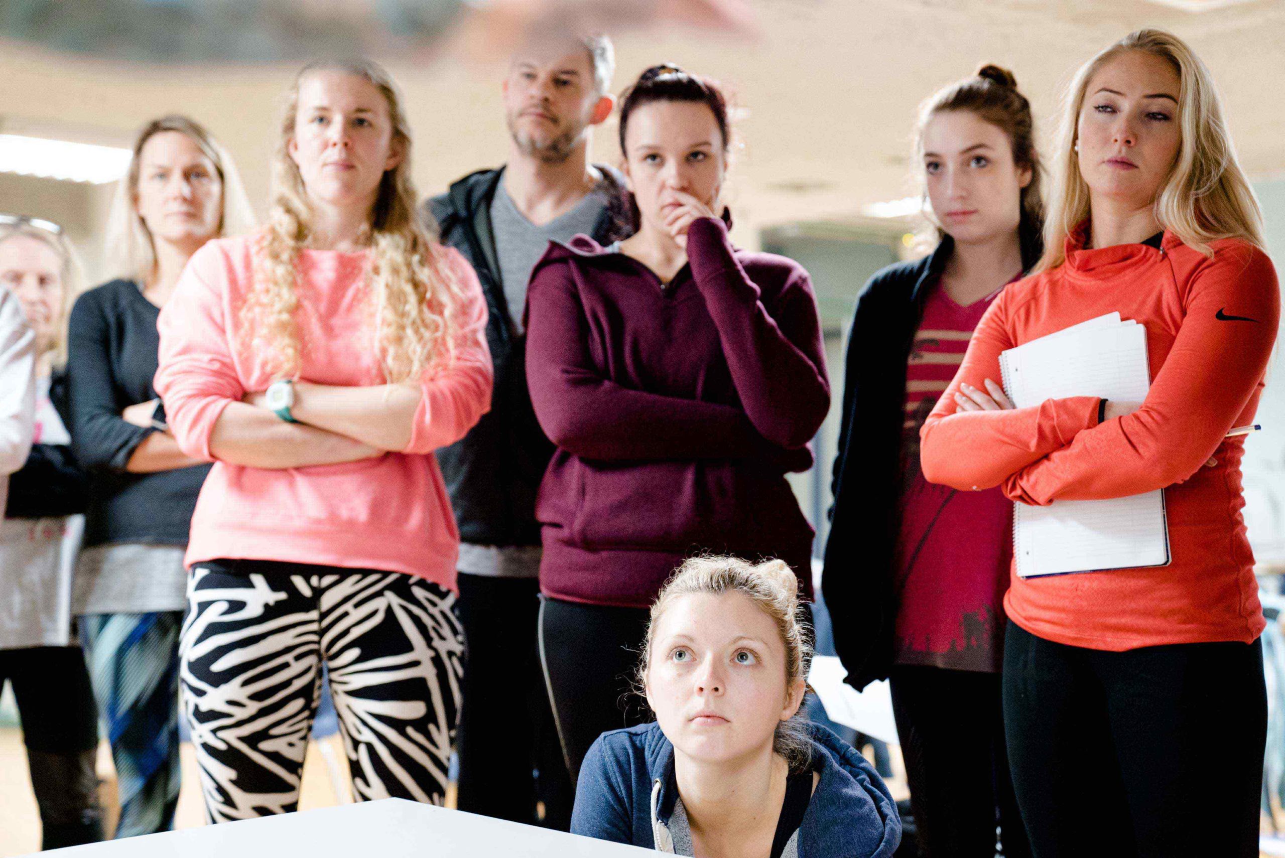 Group of people standing and one kneeling in a casual indoor setting, some with arms crossed and one holding a notebook.