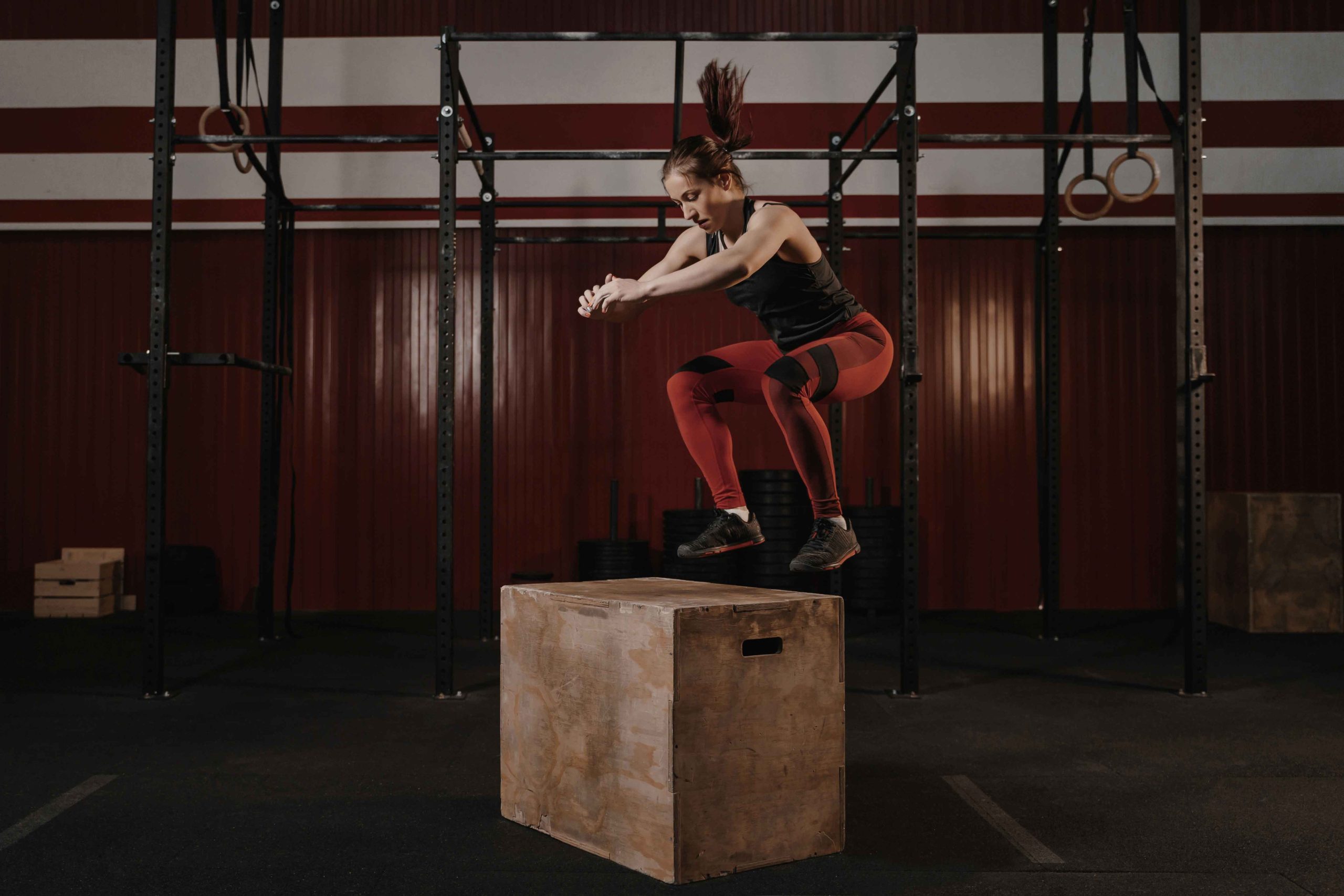 A woman in athletic wear performs a box jump in a gym. She is mid-air, displaying strength and focus against a backdrop of gym equipment.