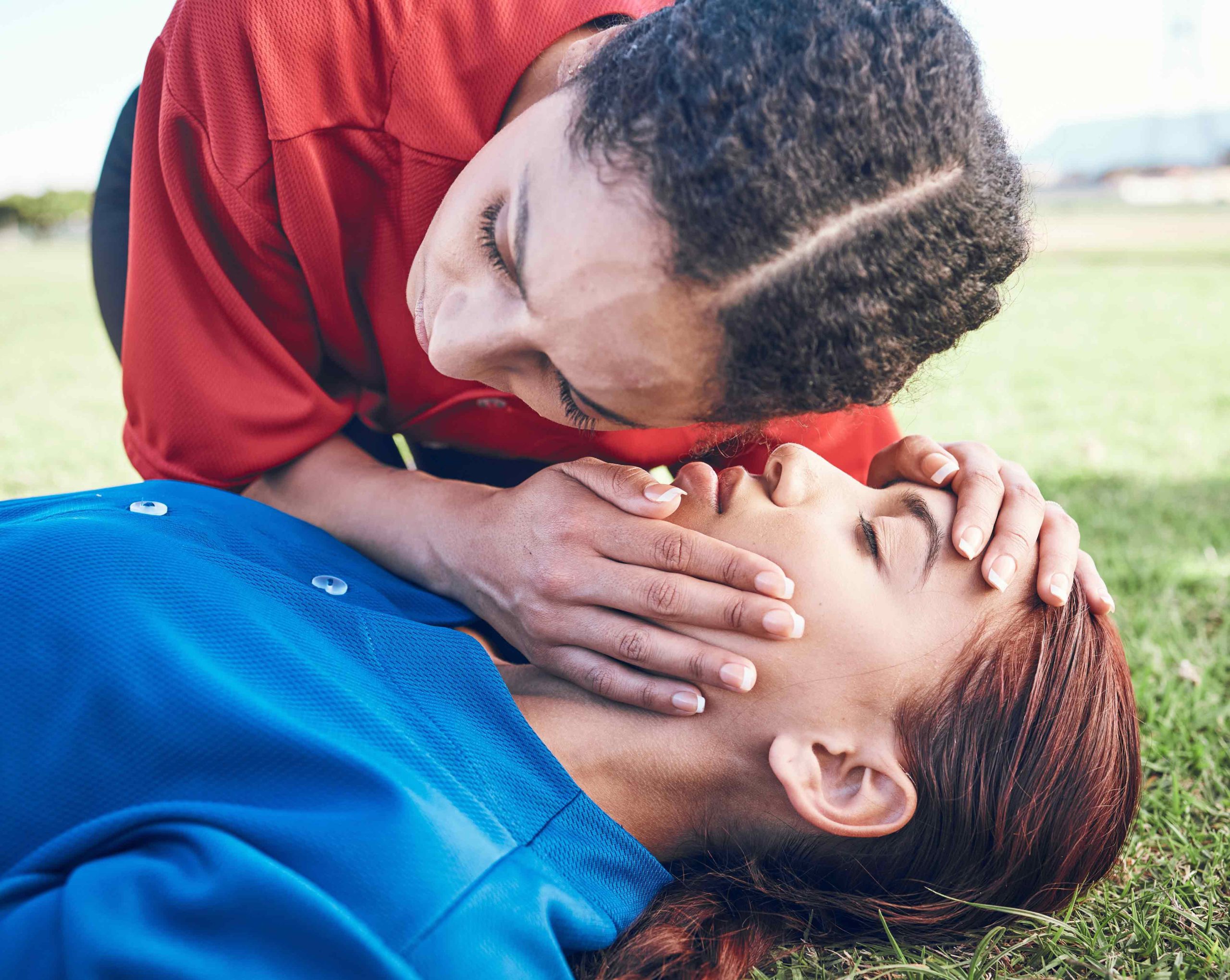 Person in red shirt performing mouth-to-mouth resuscitation on another person lying on grass, wearing a blue shirt.