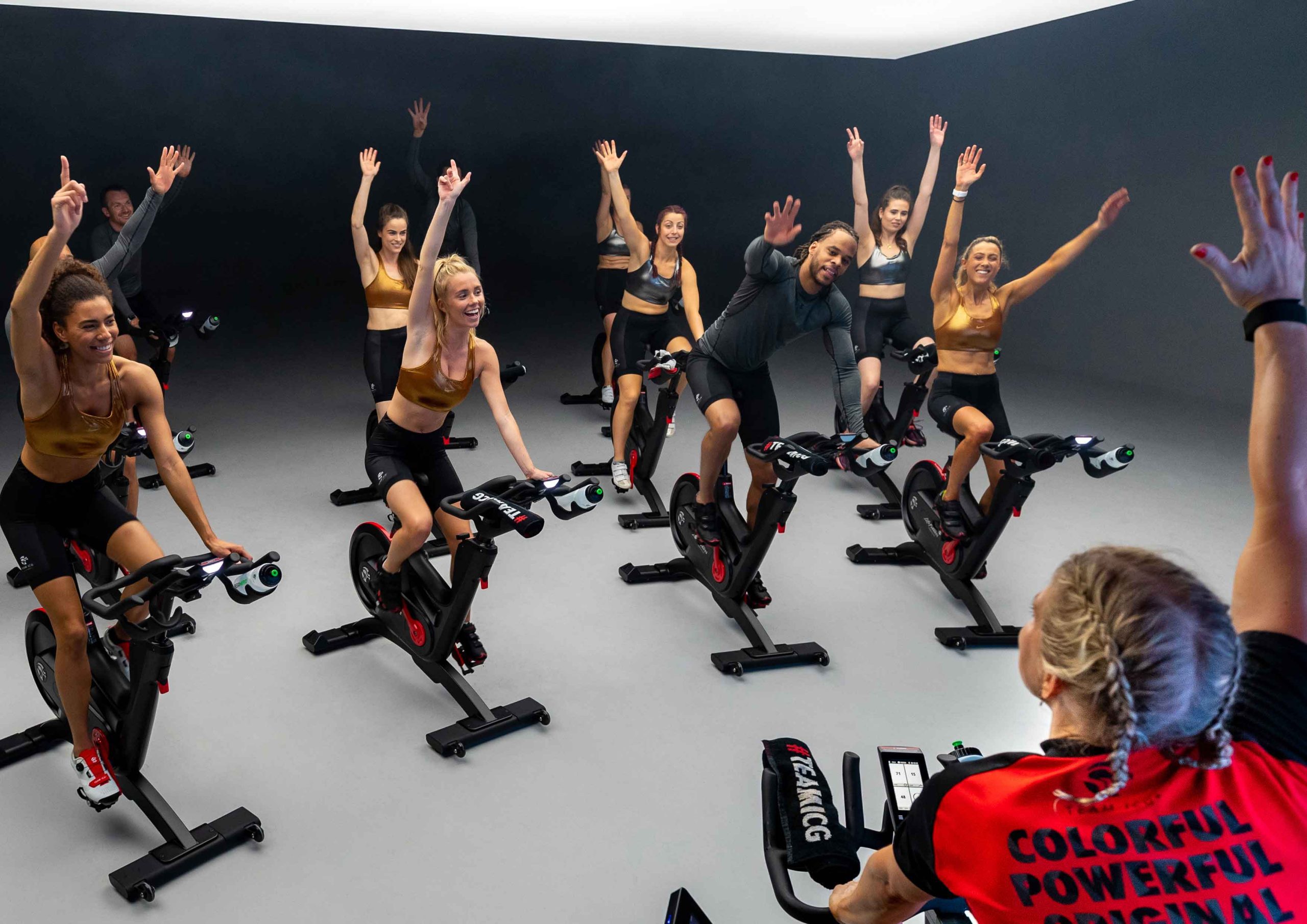 Indoor spin class with multiple participants on stationary bikes raising one arm, led by an instructor in a red and black shirt.
