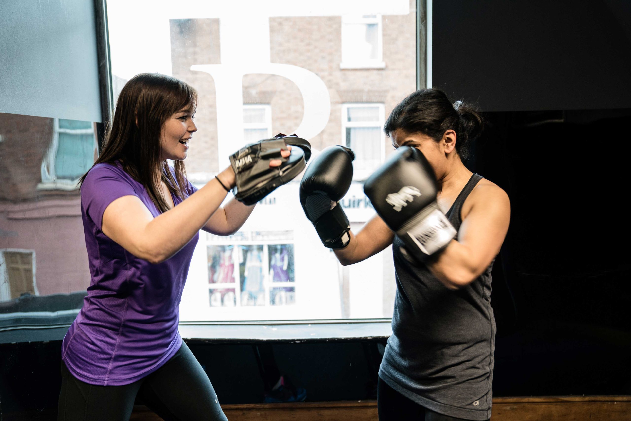 Two women sparring in a gym, one in a purple shirt holds focus mitts while the other in a black tank top wears boxing gloves, displaying focus and energy.