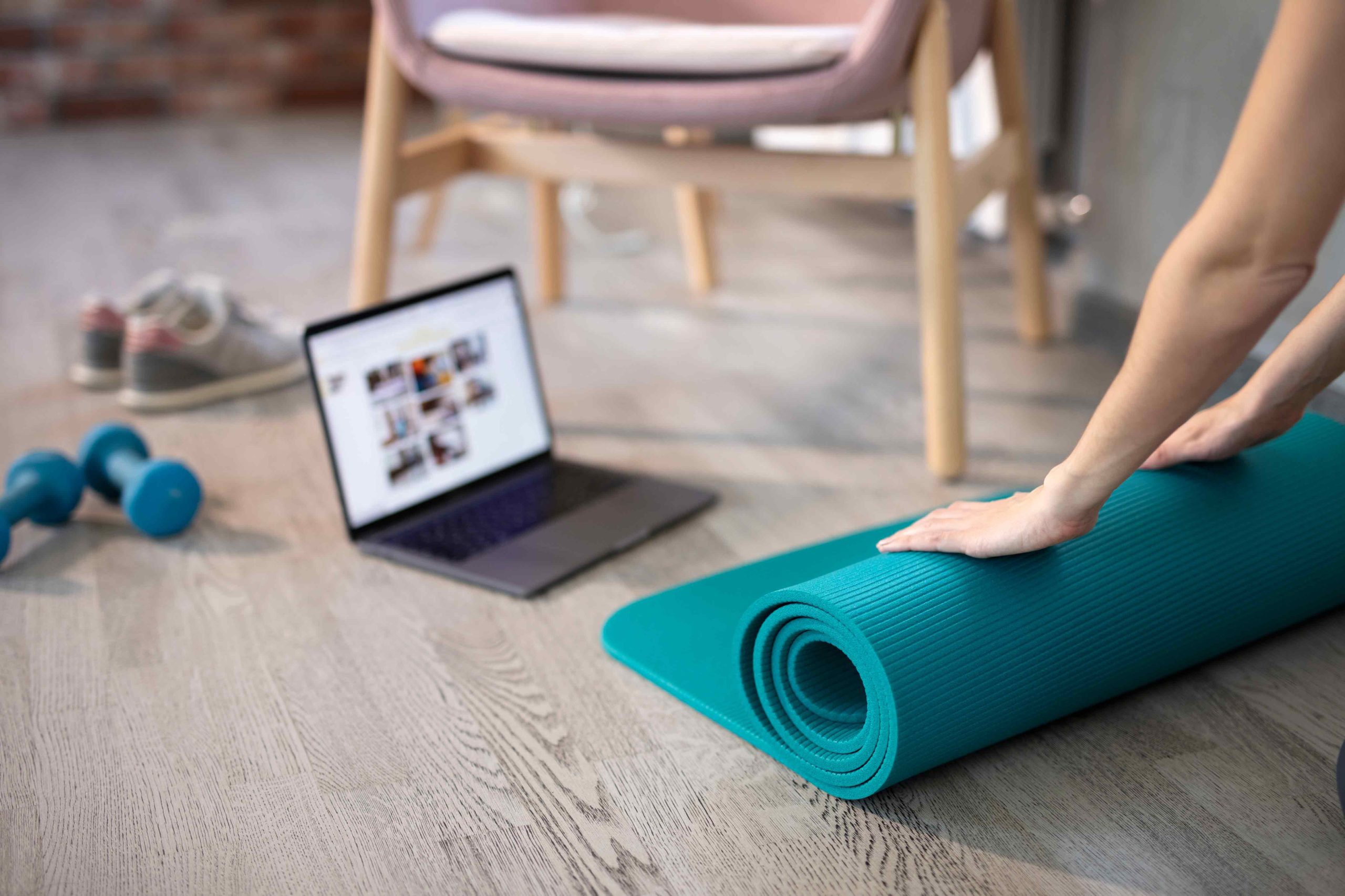 Hands rolling out a teal yoga mat on a wooden floor. Nearby are a laptop displaying fitness content, blue dumbbells, and sneakers. The setting is calm.