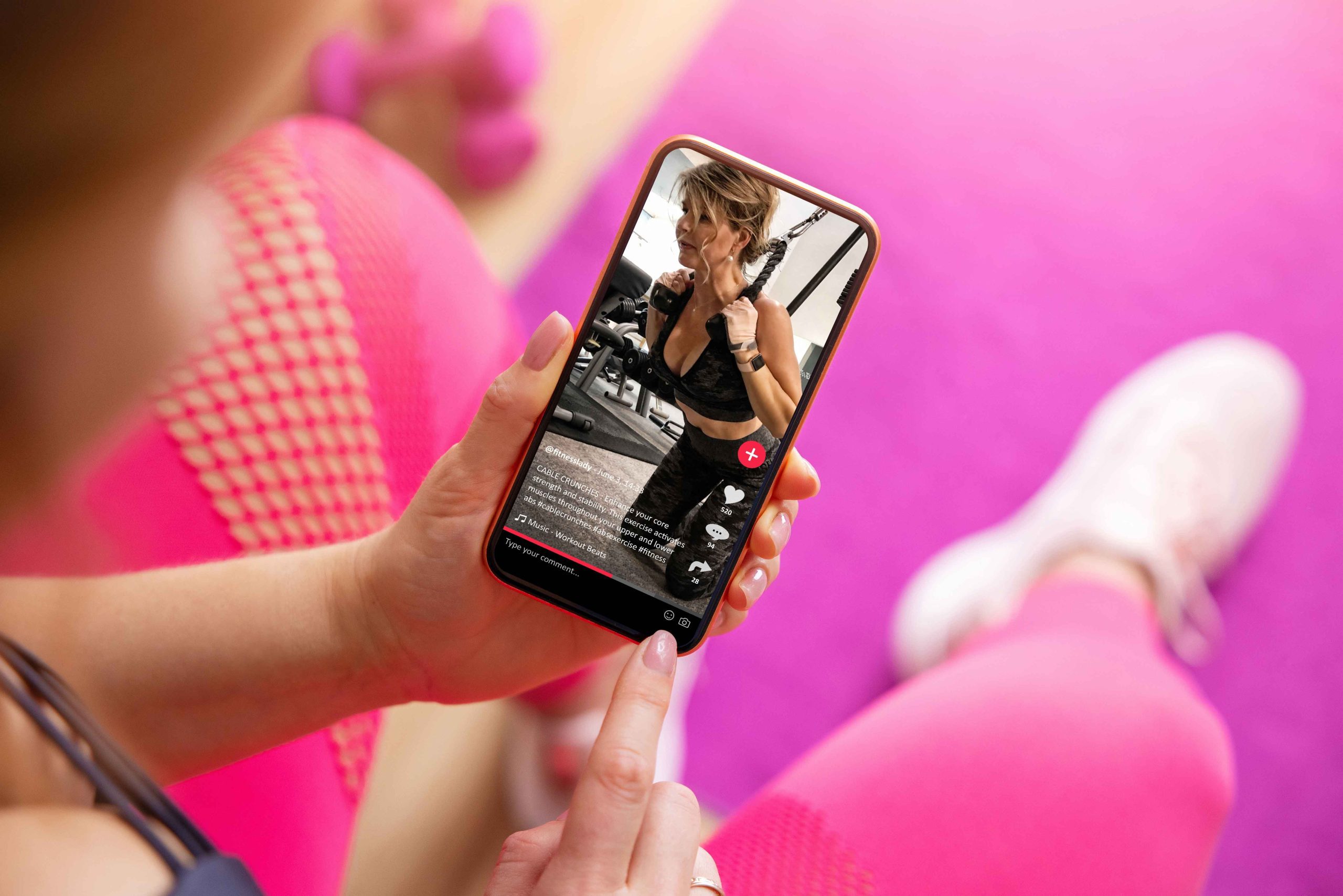 Person in pink leggings watches a fitness workout video on a smartphone while sitting on a pink exercise mat.