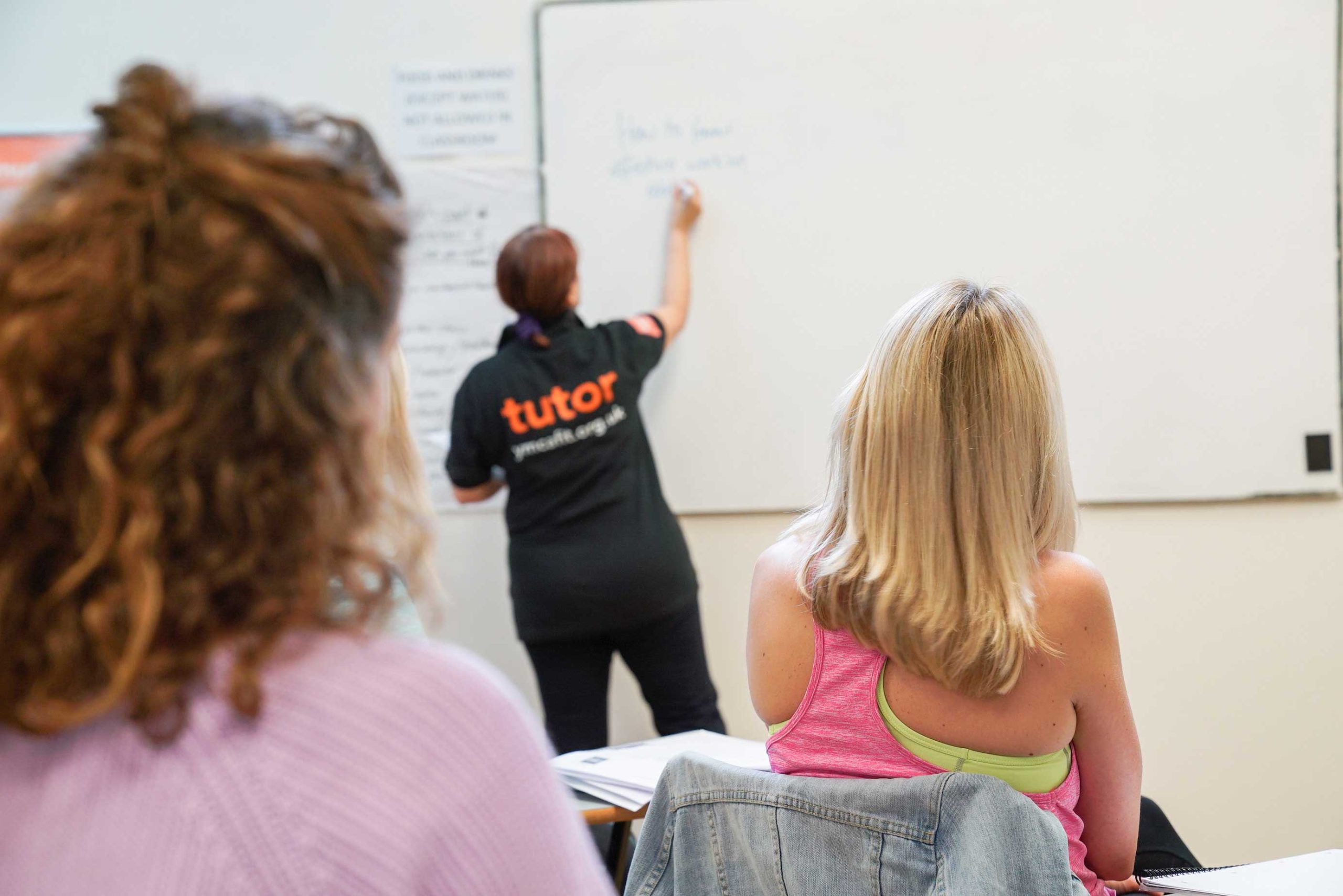 Classroom scene with a tutor writing on a whiteboard and two students seated, facing the board, one wearing a pink top.