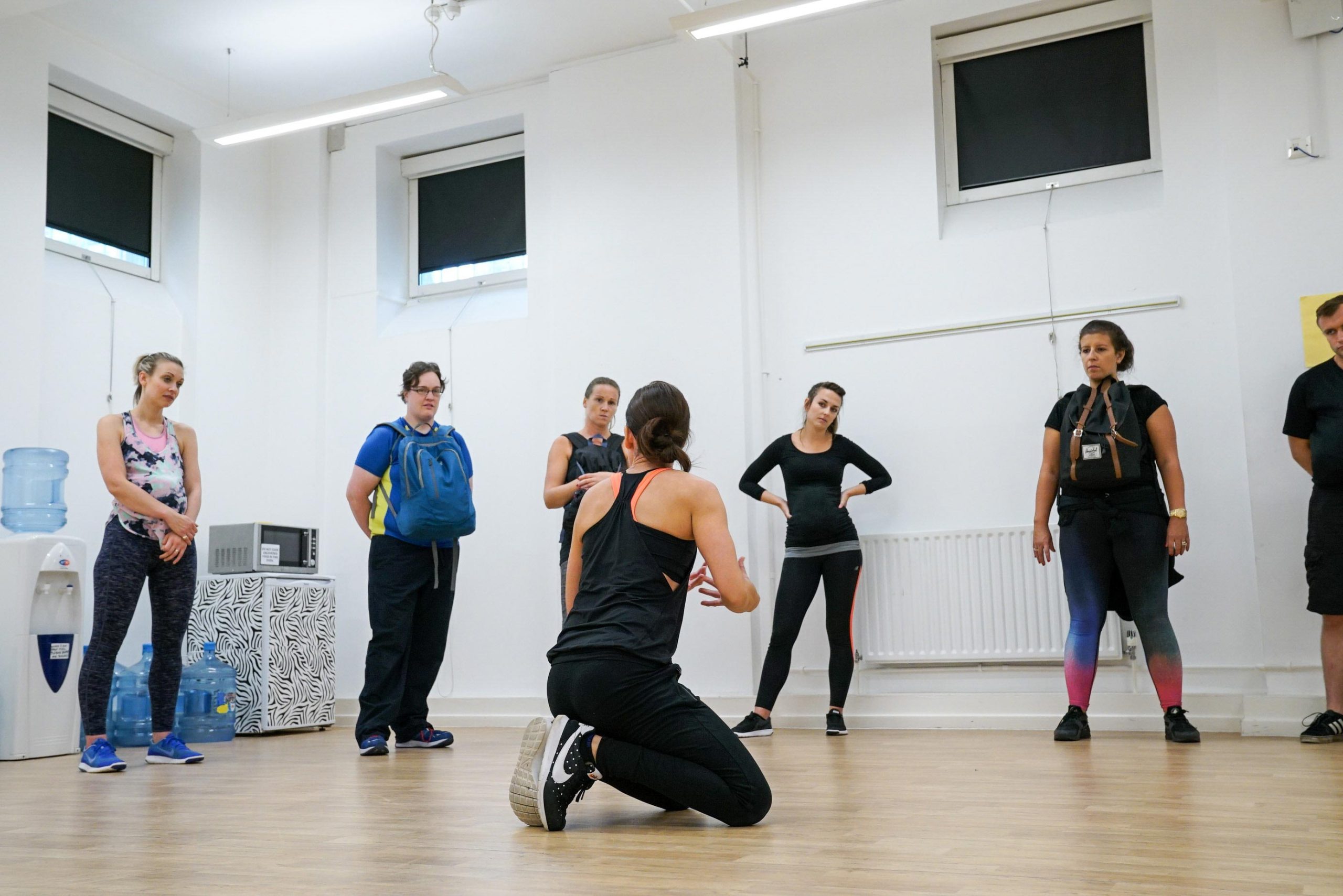 Group fitness instructor kneels on floor demonstrating exercise to standing participants in a bright studio room.