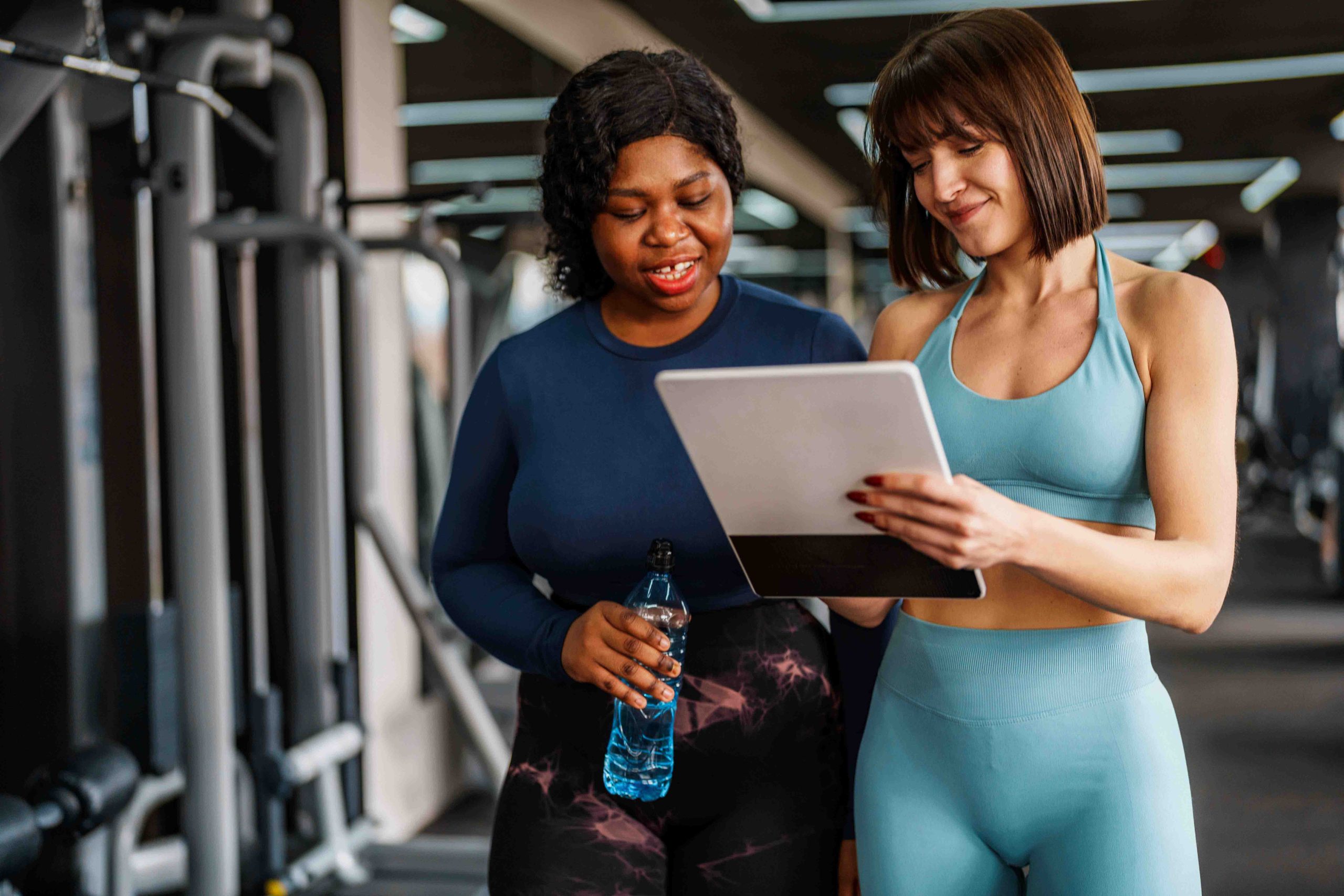 Two women in workout clothes at a gym, one holding a water bottle and the other holding a tablet.