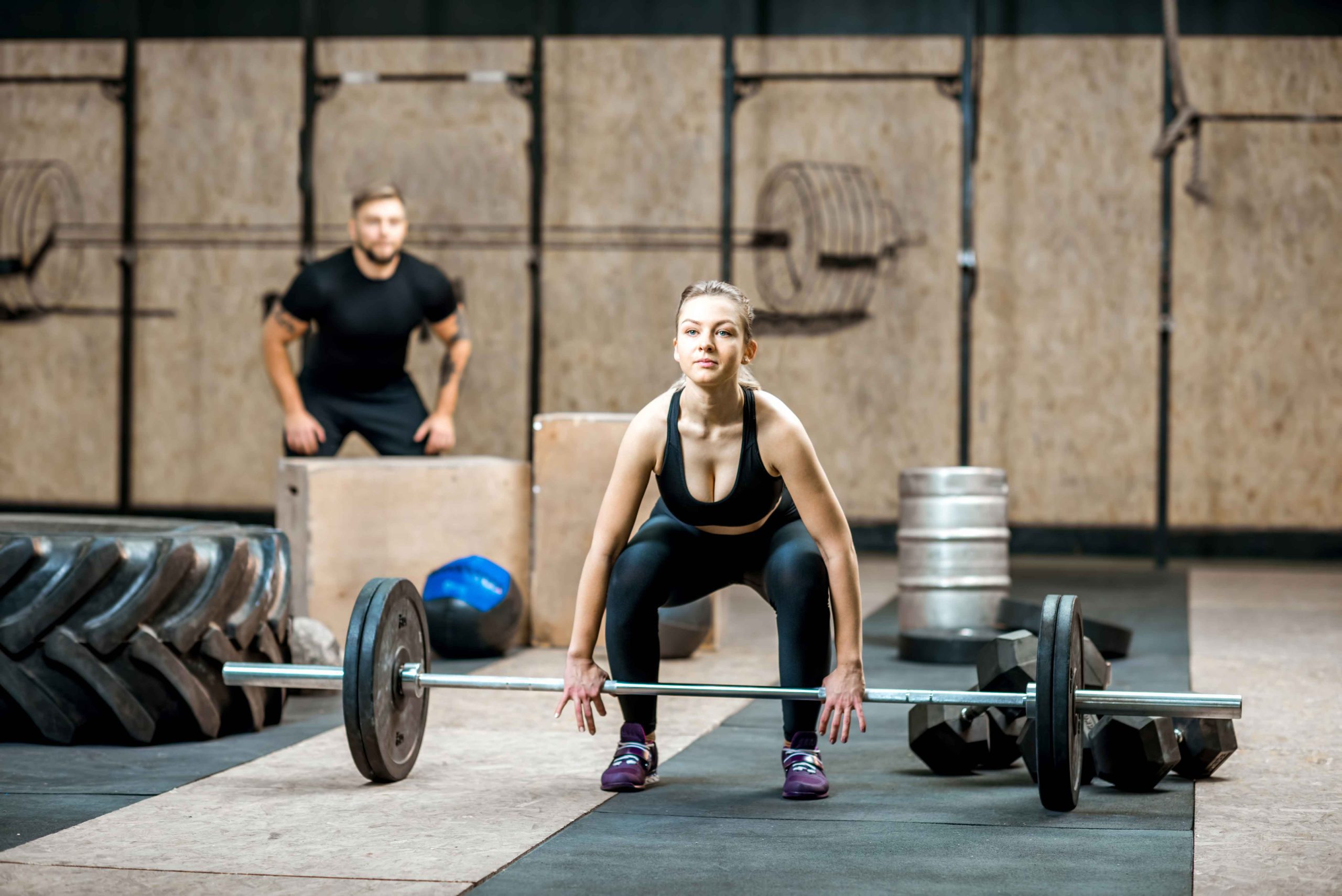 Woman lifting barbell in a gym with a trainer watching, surrounded by fitness equipment like tires, weights, and boxes.