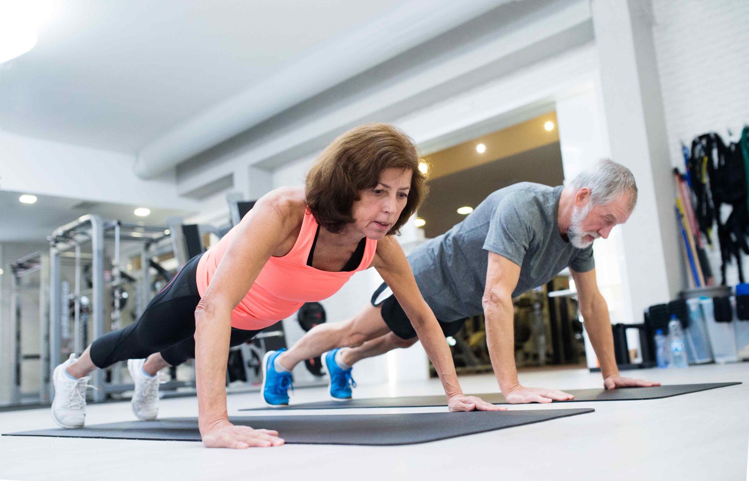Two older adults doing push-ups on mats in a bright gym with fitness equipment in the background.