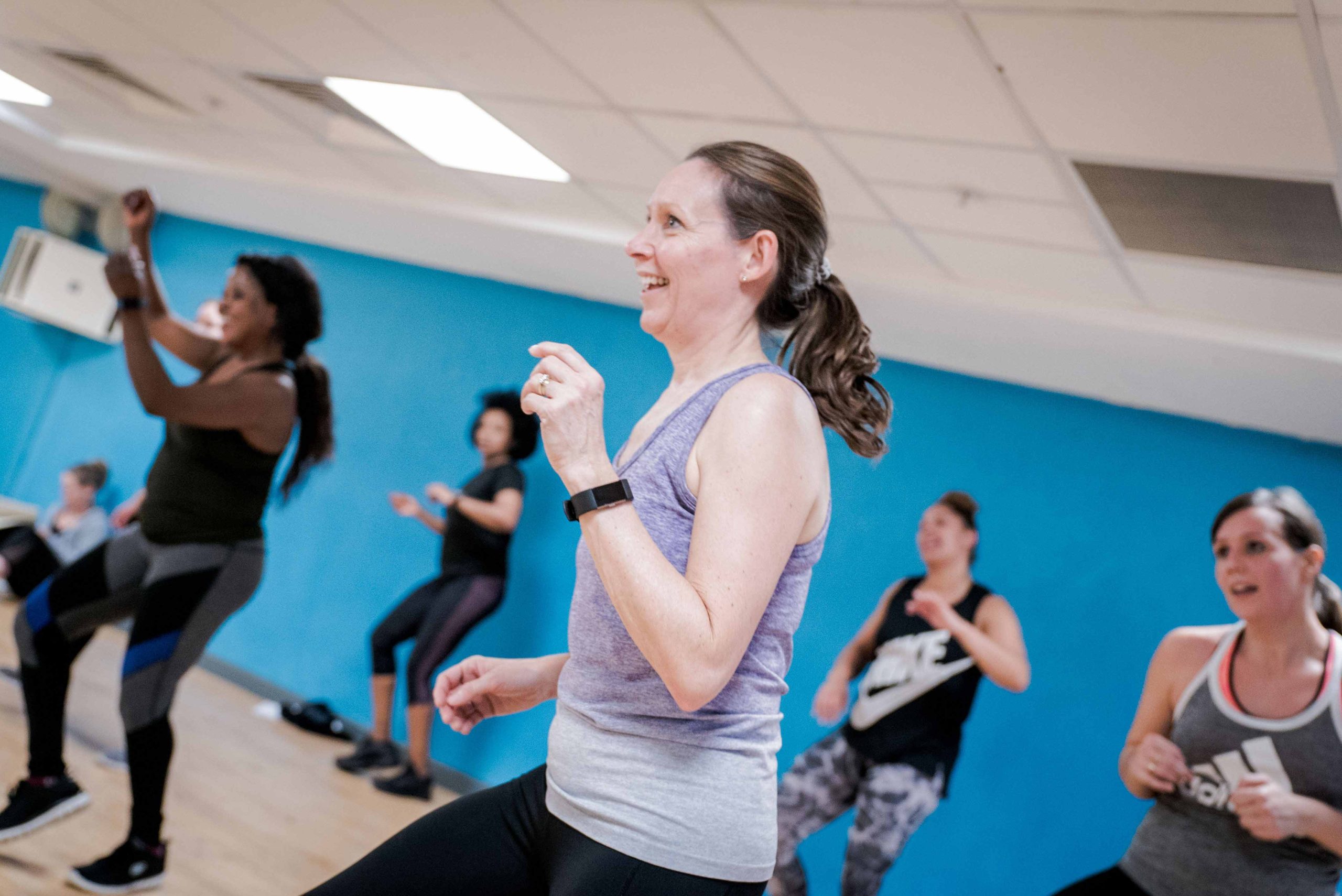 Group of people participating in a fitness class, performing aerobic exercises in a bright gym with blue walls.