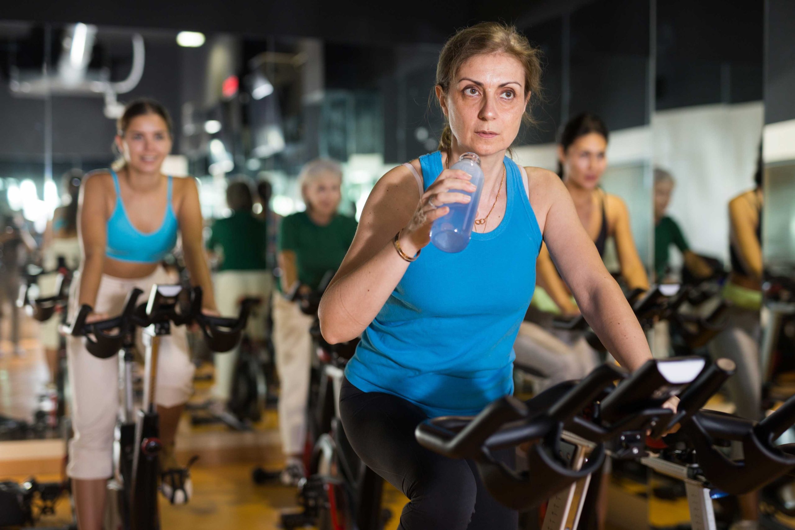 Group of women cycling indoors in a spin class, with one woman in a blue top drinking water from a bottle.