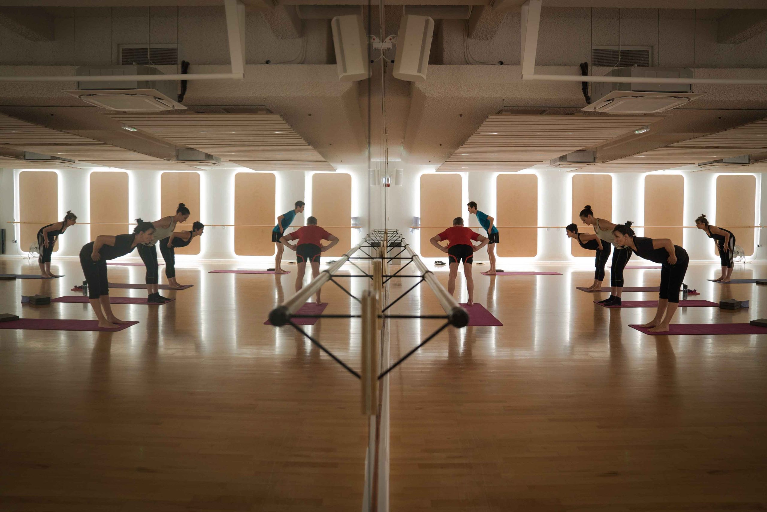 A yoga class with people stretching in a mirrored studio. Sunlit windows illuminate the wooden floor, creating a calm, focused atmosphere.