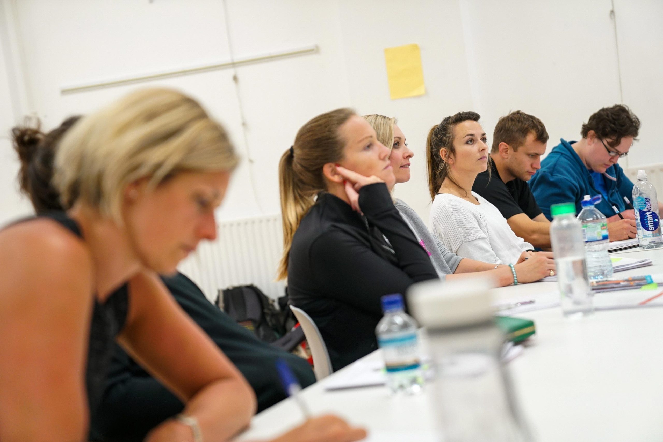 A group of people sitting at a table in a classroom or meeting setting, engaged in writing or listening attentively.
