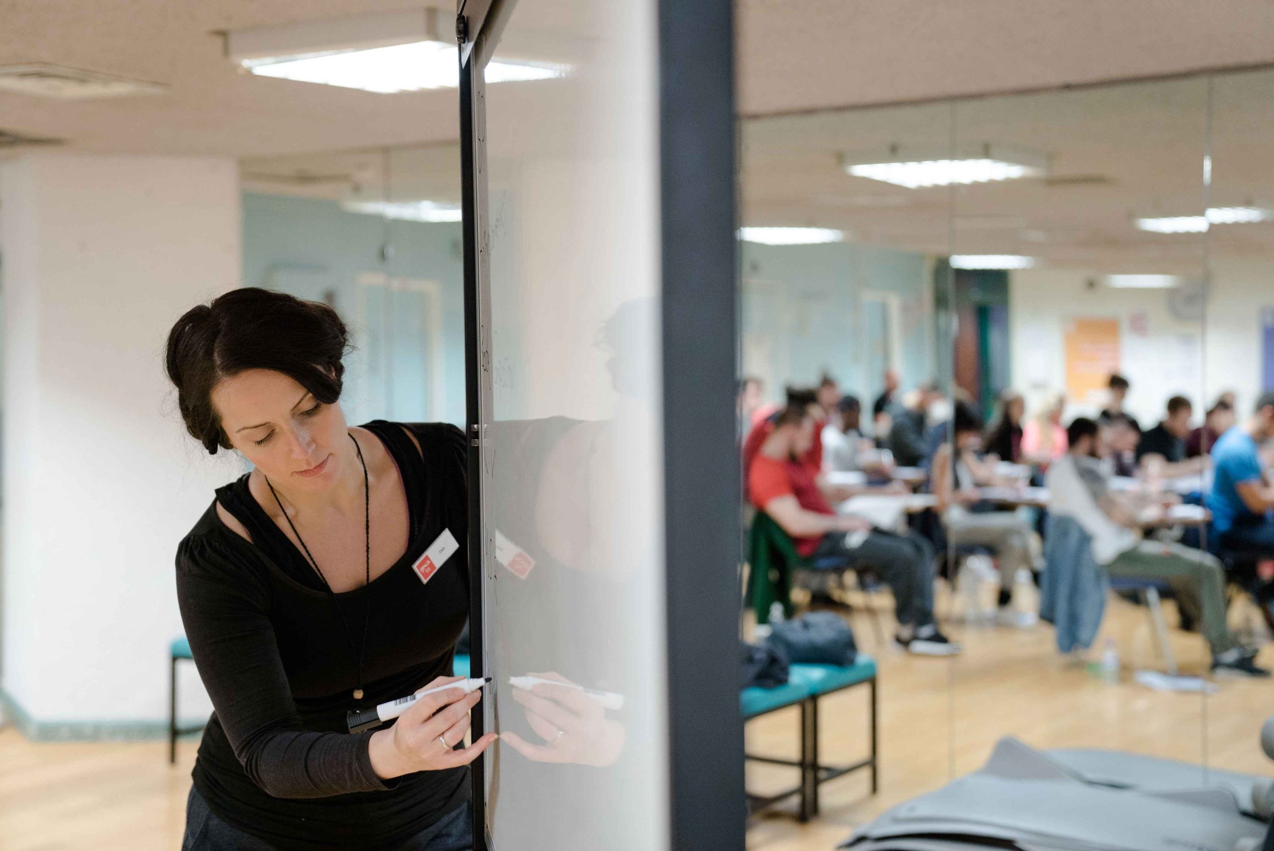Teacher writing on a whiteboard with students sitting and listening in a classroom with wooden floors and bright lighting.