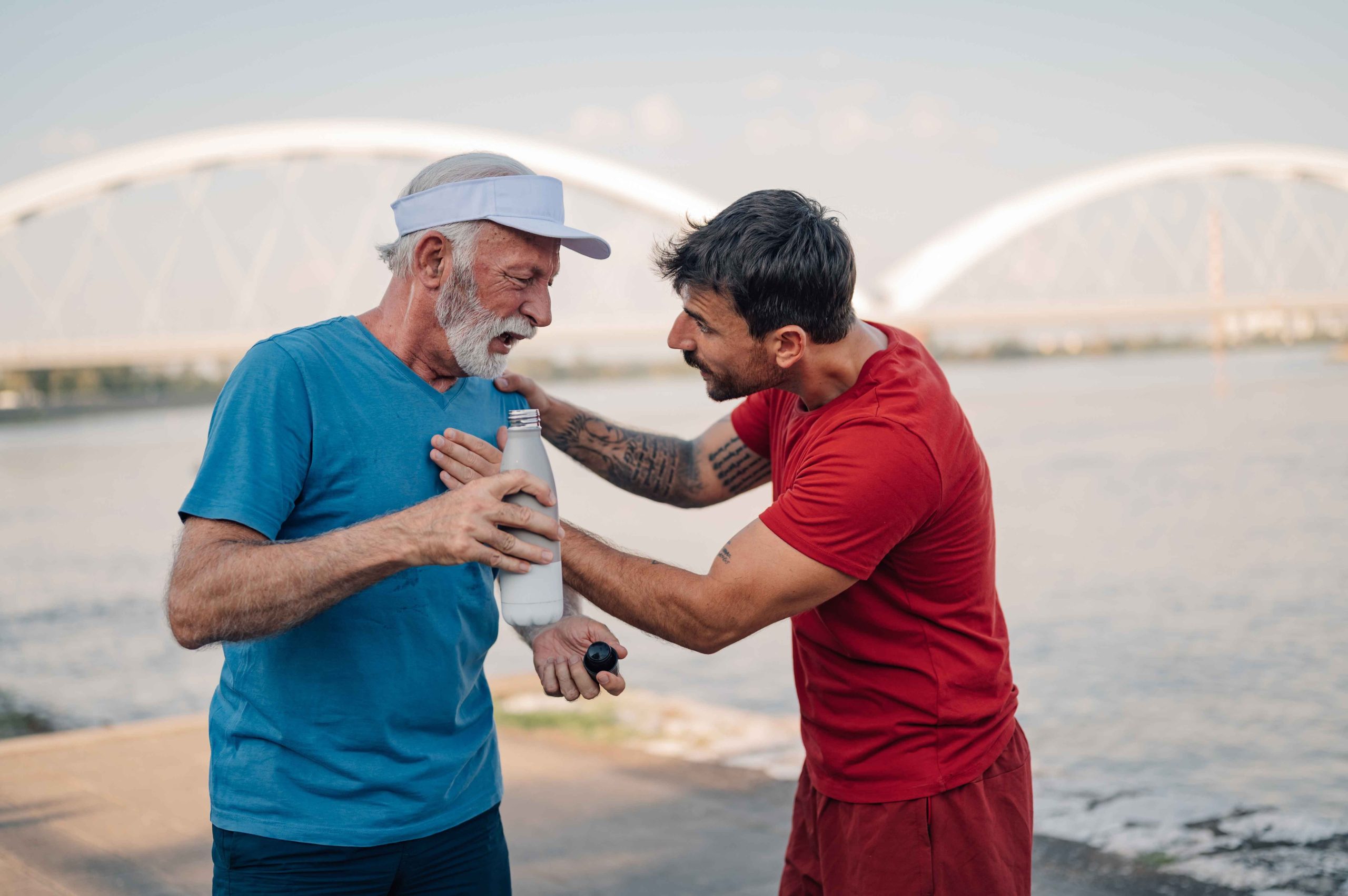 Two men outdoors by a river, one in a red shirt helping the other in a blue shirt who appears to be in discomfort.