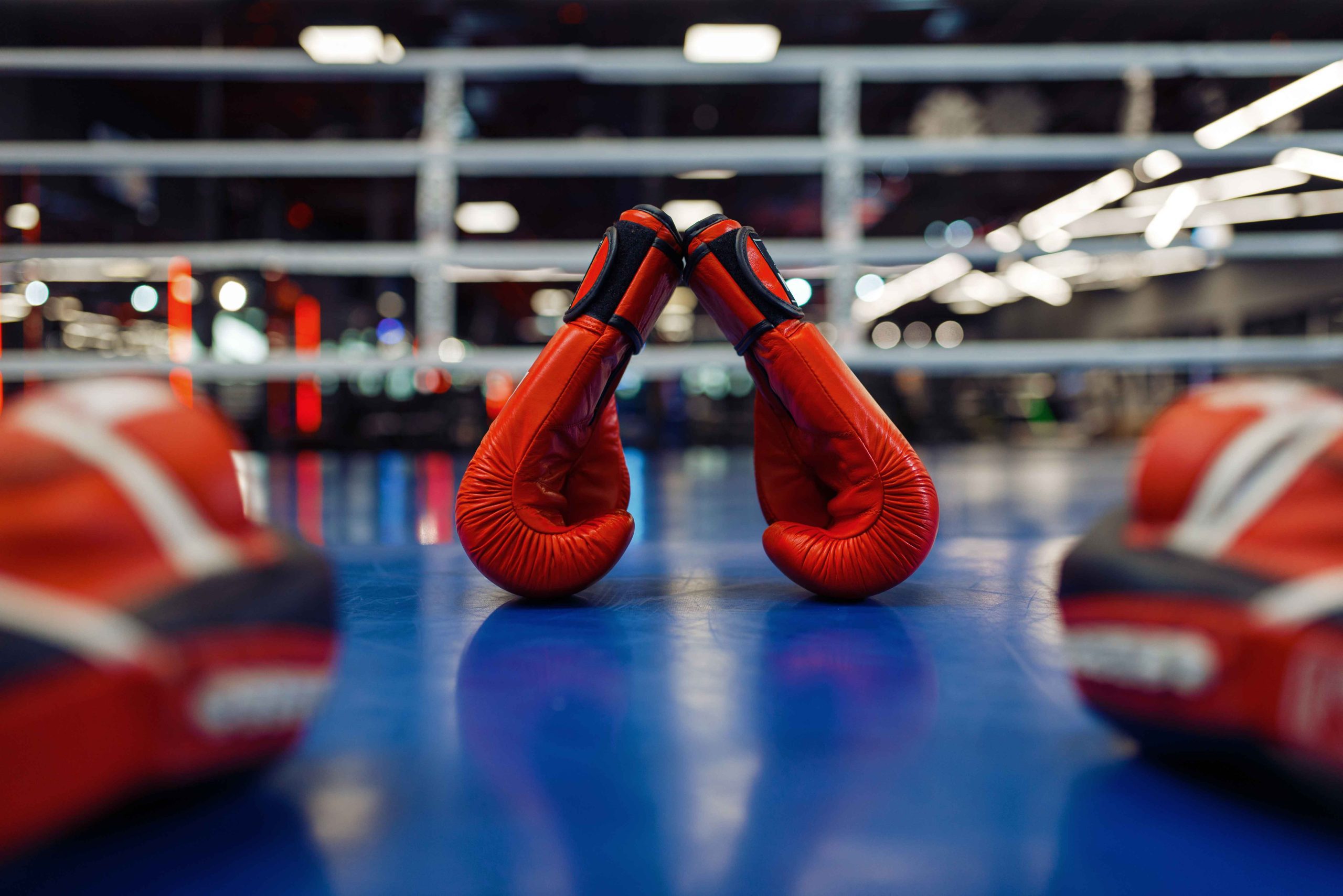 Red boxing gloves rest upright on a blue mat in a gym. The focus is on the gloves, with blurred gym equipment and rings in the background, creating a dynamic and energetic atmosphere.