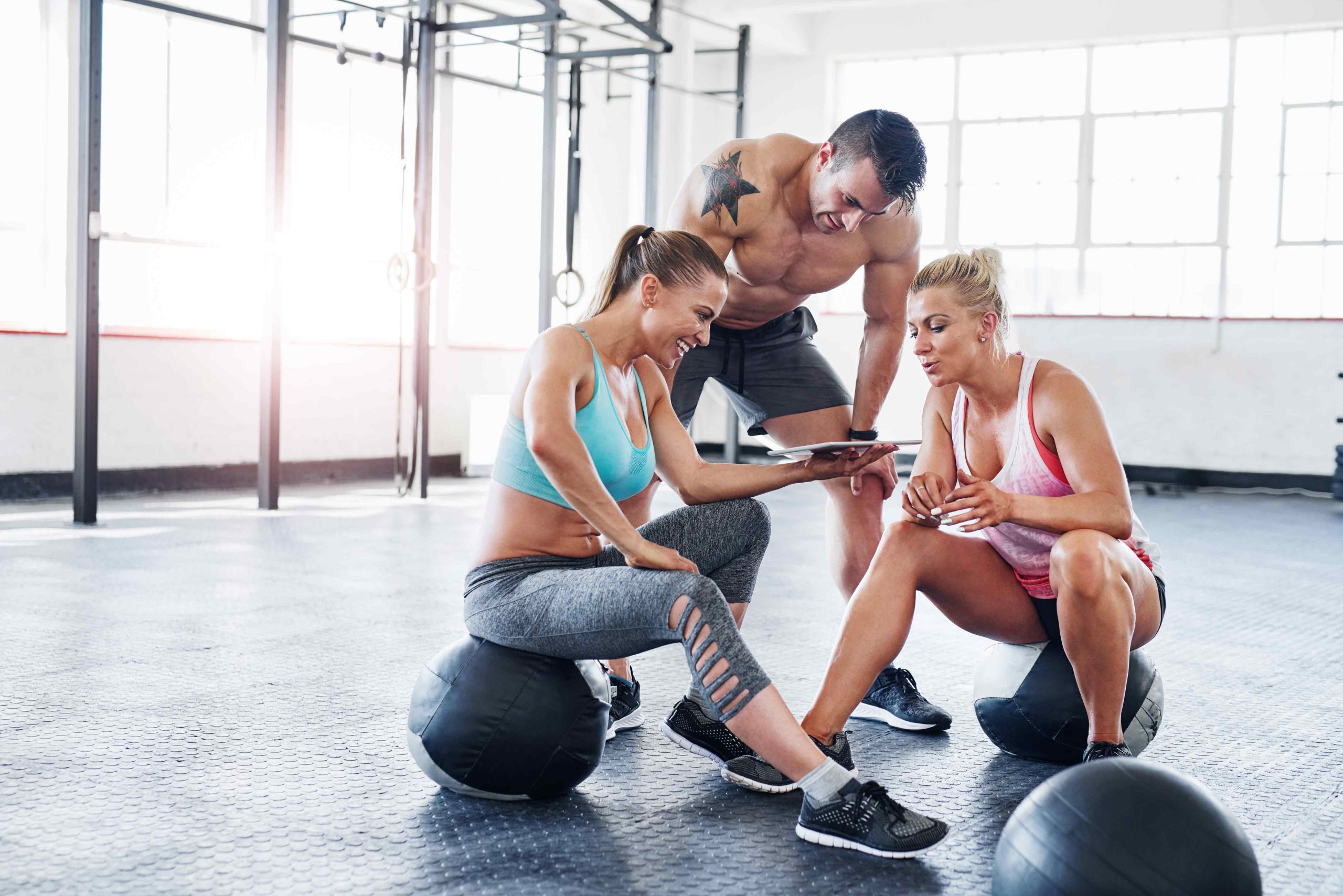 Three fit people in workout clothes using a tablet and sitting on exercise balls inside a gym with large windows.