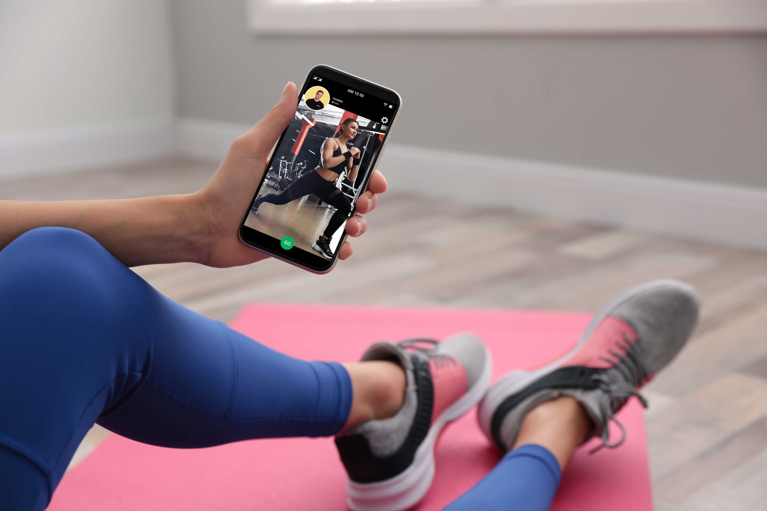 Person in workout attire sitting on a pink mat holding a phone showing a fitness instructor doing lunges in a gym.