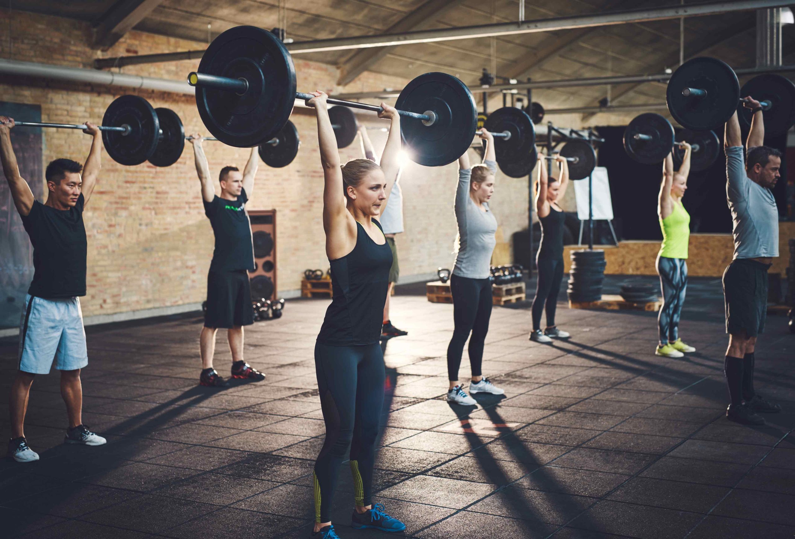 A group of six people in athletic wear lift barbells overhead in a gym, displaying strength and determination. The room has high ceilings and a motivational vibe.