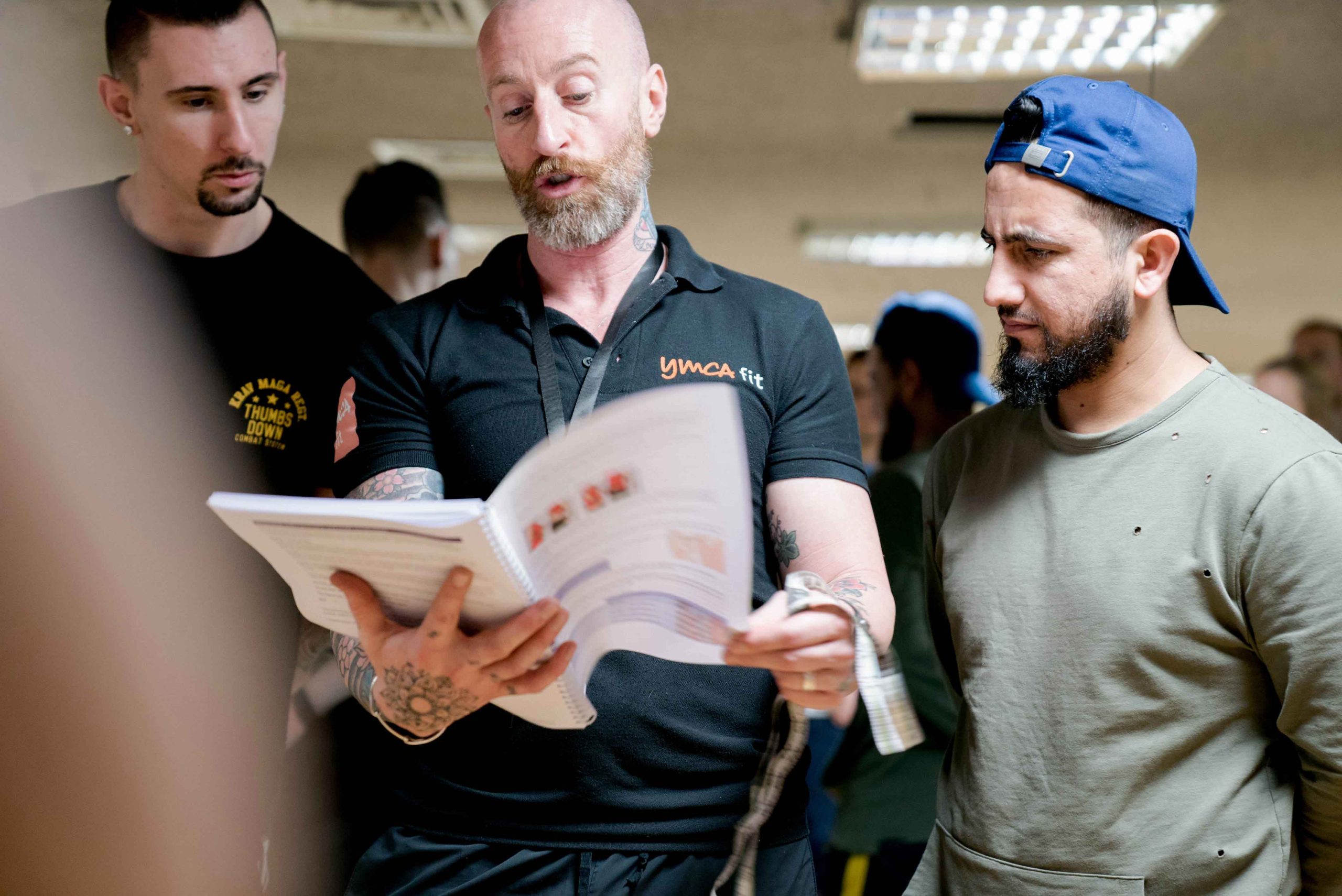 Three men in casual clothing discuss while one reads from a booklet in a crowded indoor setting.