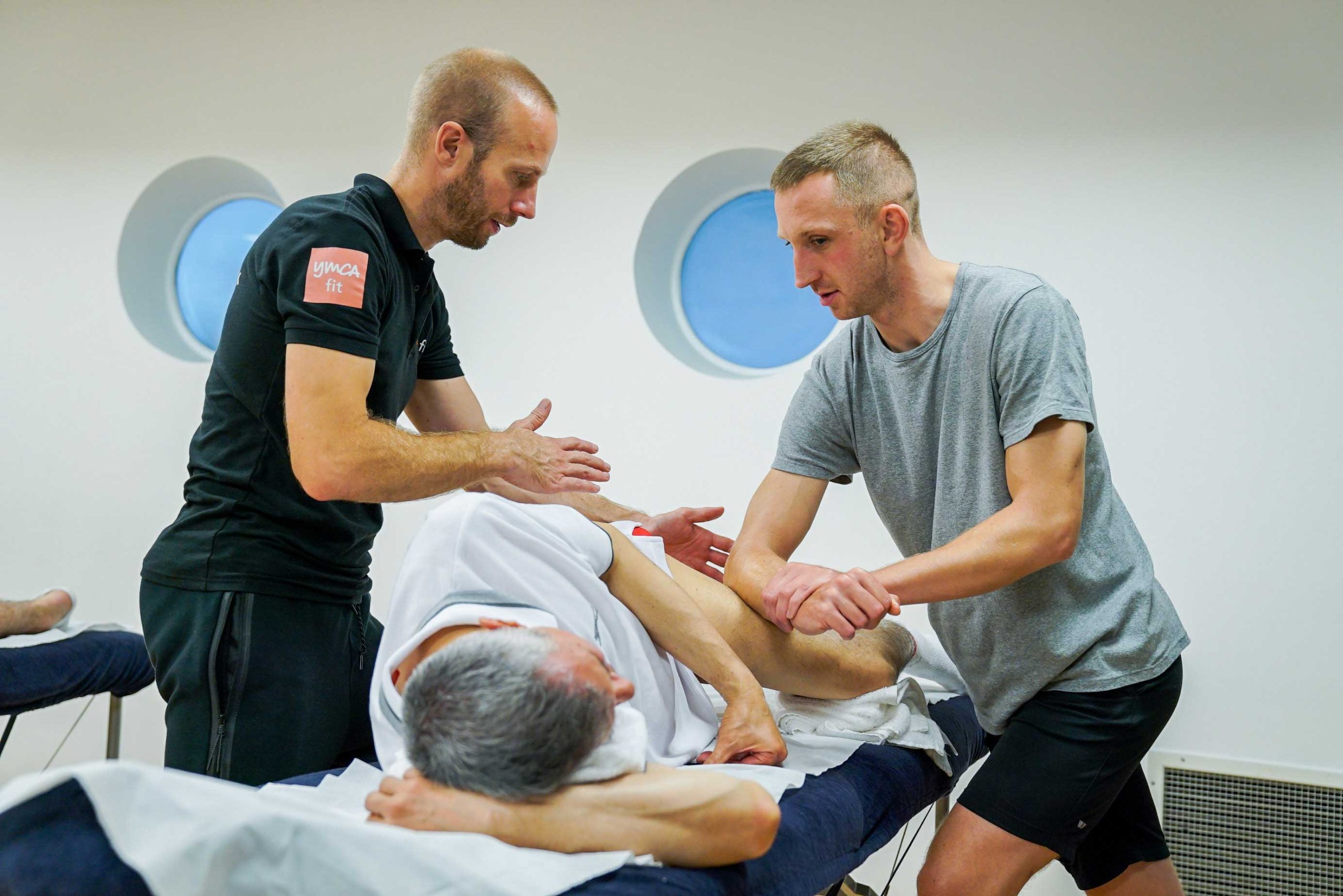 Two therapists assisting a male patient lying on a treatment table in a bright room with round windows.