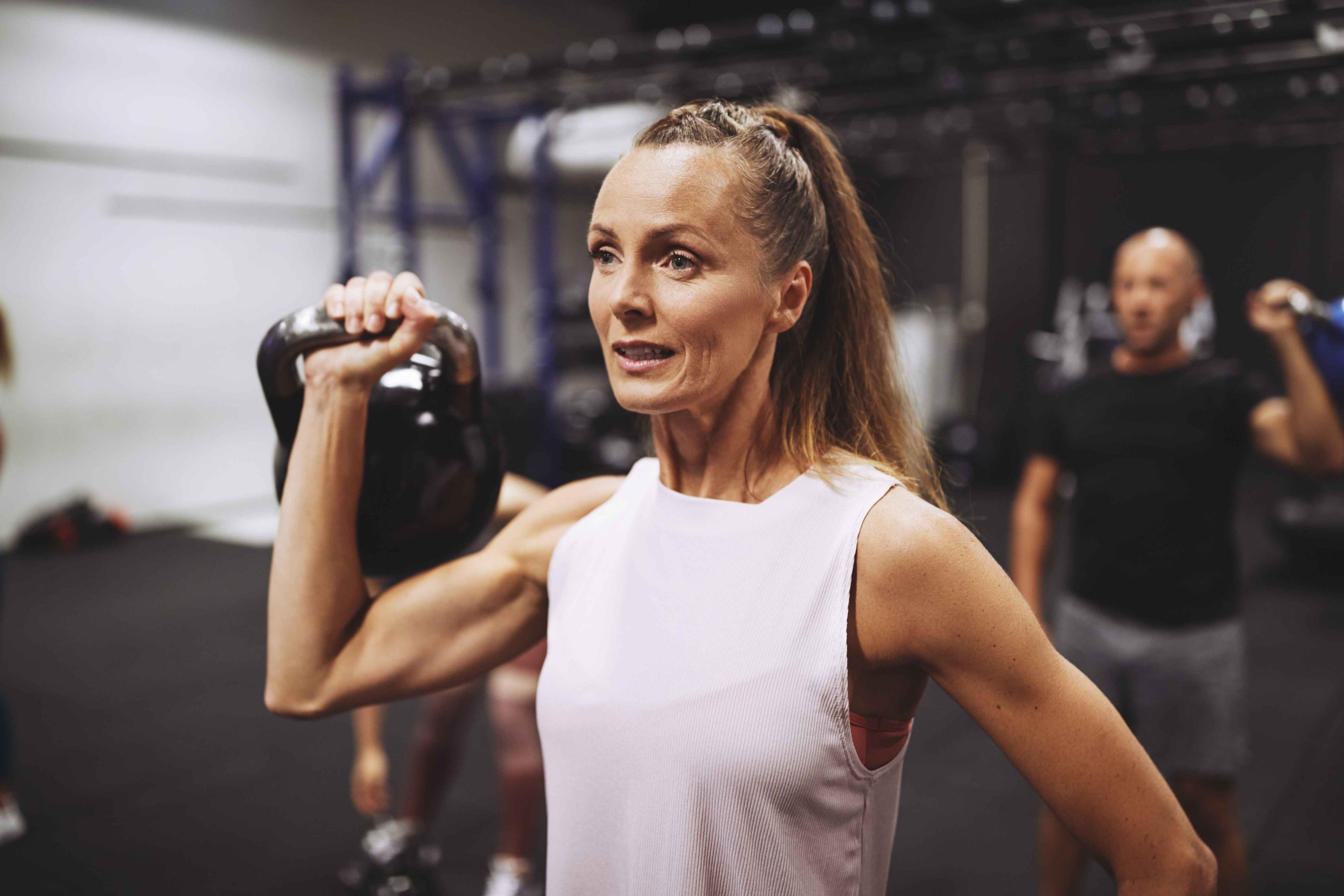 Woman lifting a kettlebell in a gym with other people exercising in the background.