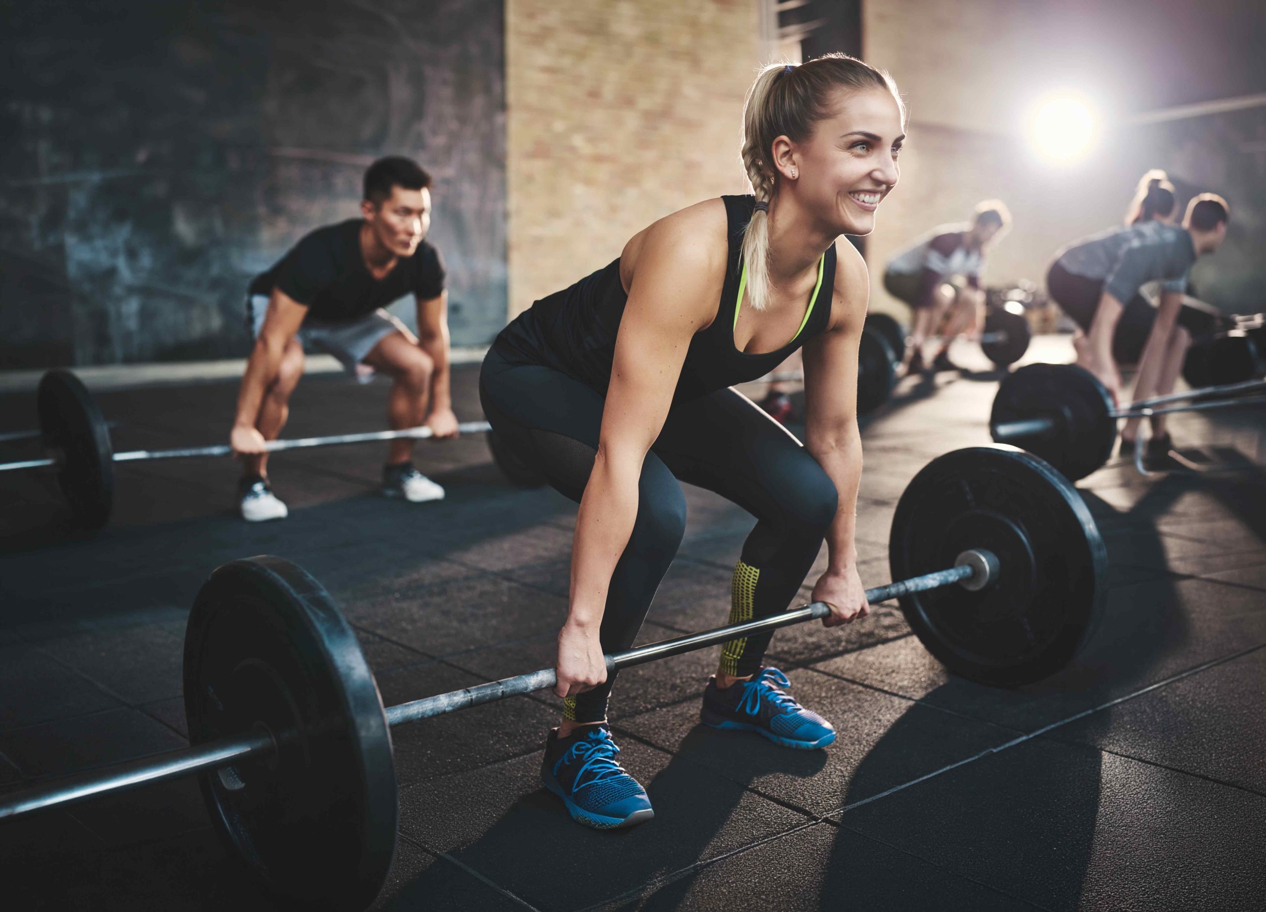 Group of people lifting barbells during a workout session in a gym with bright lighting and rubber flooring.