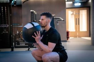 A man in a gym performs a squat while holding a medicine ball at chest height. He wears black athletic clothing, appearing focused and determined.