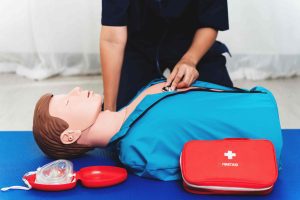 Person practicing CPR and using a stethoscope on a training dummy, with a first aid kit and mask nearby on a blue mat.