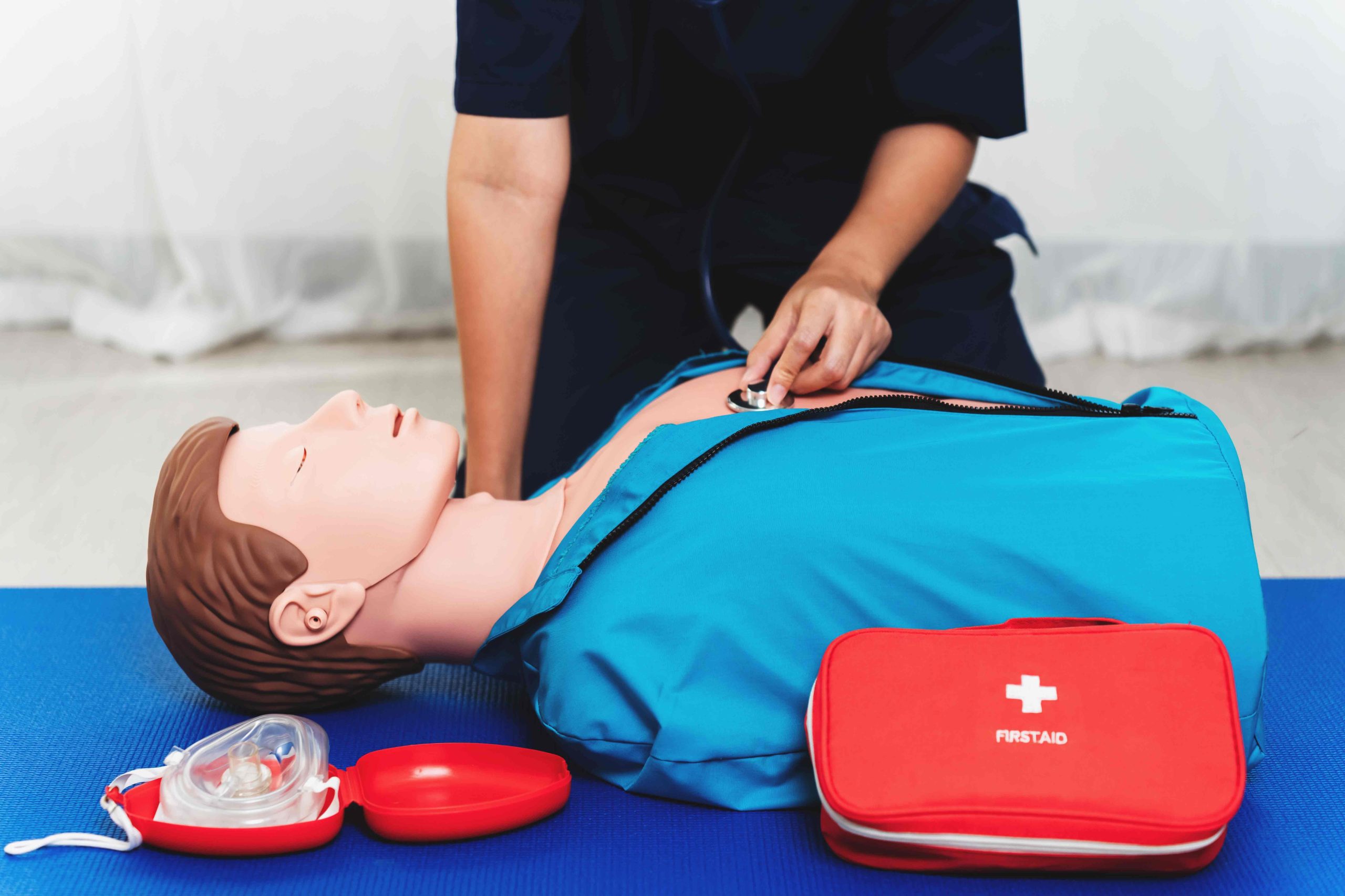 Person practicing CPR and using a stethoscope on a training dummy, with a first aid kit and mask nearby on a blue mat.