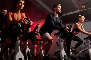 Group of people exercising on stationary bikes in a dimly lit indoor cycling studio with red and blue accent lighting.