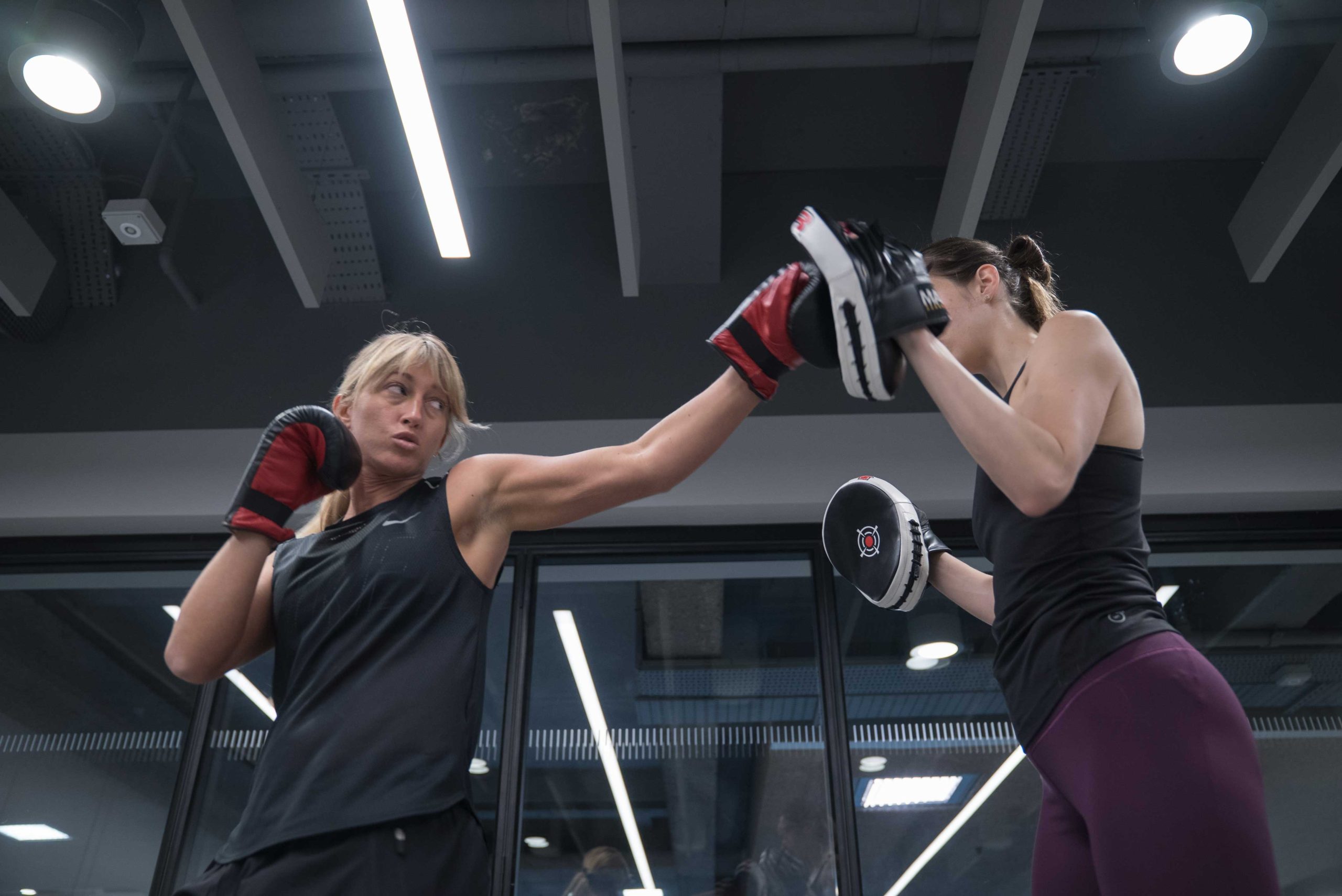 Two women practice boxing in a gym. The woman on the left throws a punch, while the one on the right holds up pads. The scene conveys focus and intensity.