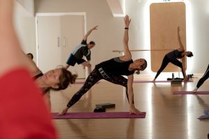 A yoga class with several participants stretching in triangle poses on purple mats in a well-lit studio. The mood is focused and serene.