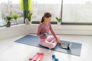 A woman in workout clothes trains with dumbbells on a yoga mat in a bright room, looking at a laptop. Exercise bands and plants surround her.
