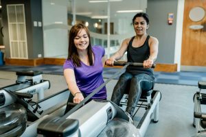 Two women exercising together on rowing machines in a gym with mirrors and wooden accents.