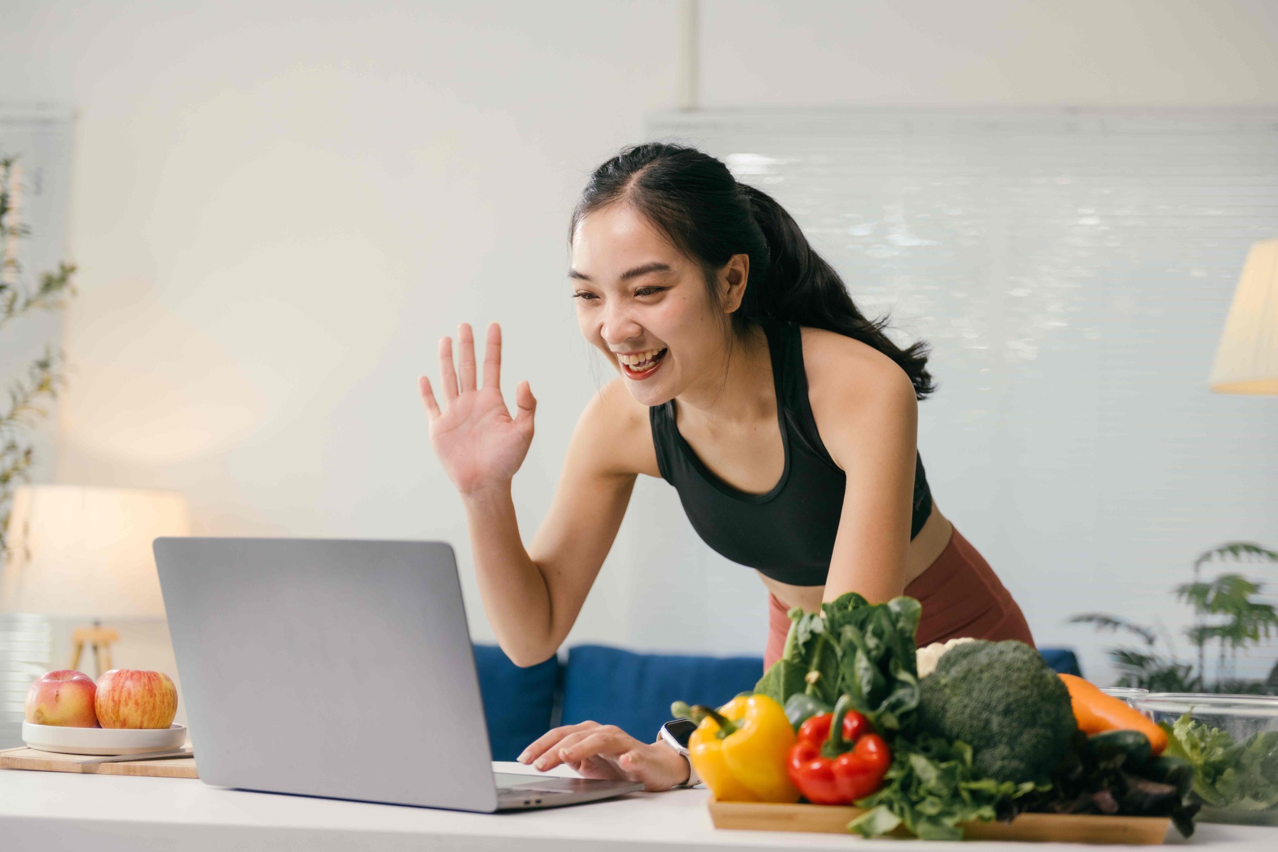 Person in workout clothes waves at laptop while surrounded by fresh vegetables on a kitchen counter.