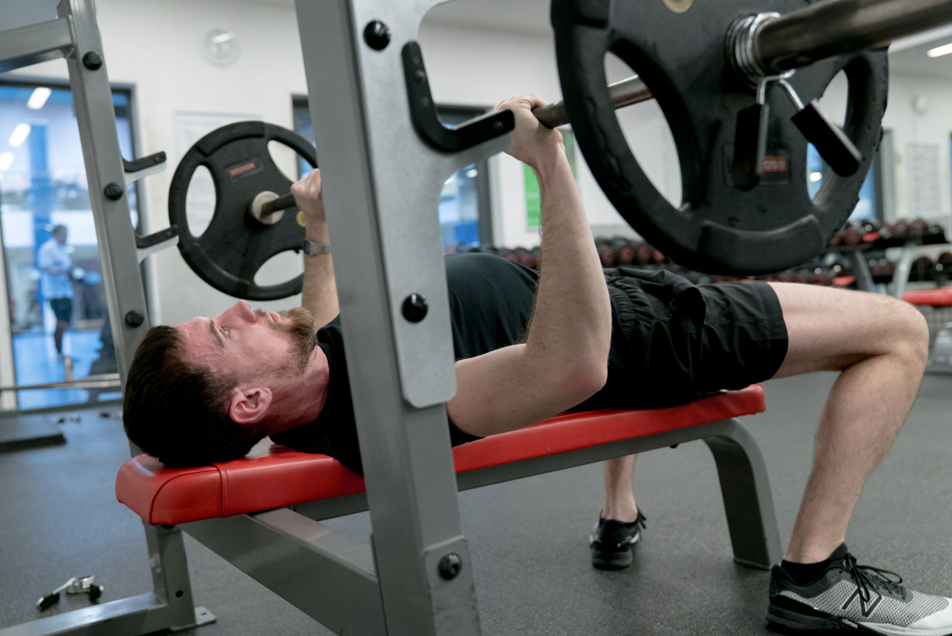 Man lifting barbell on a bench press in a gym with weight plates, wearing black shorts and gray sneakers.