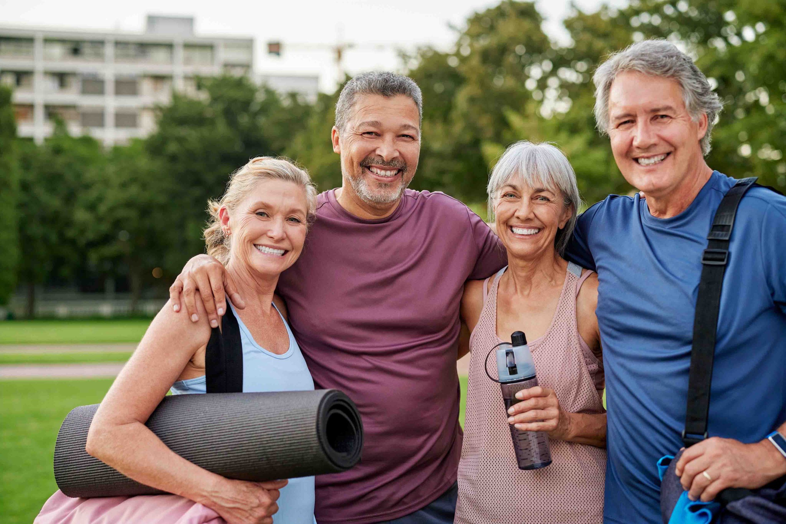 Group of four mature adults in sportswear outdoors, carrying yoga mat and water bottle, posing with arms around each other.