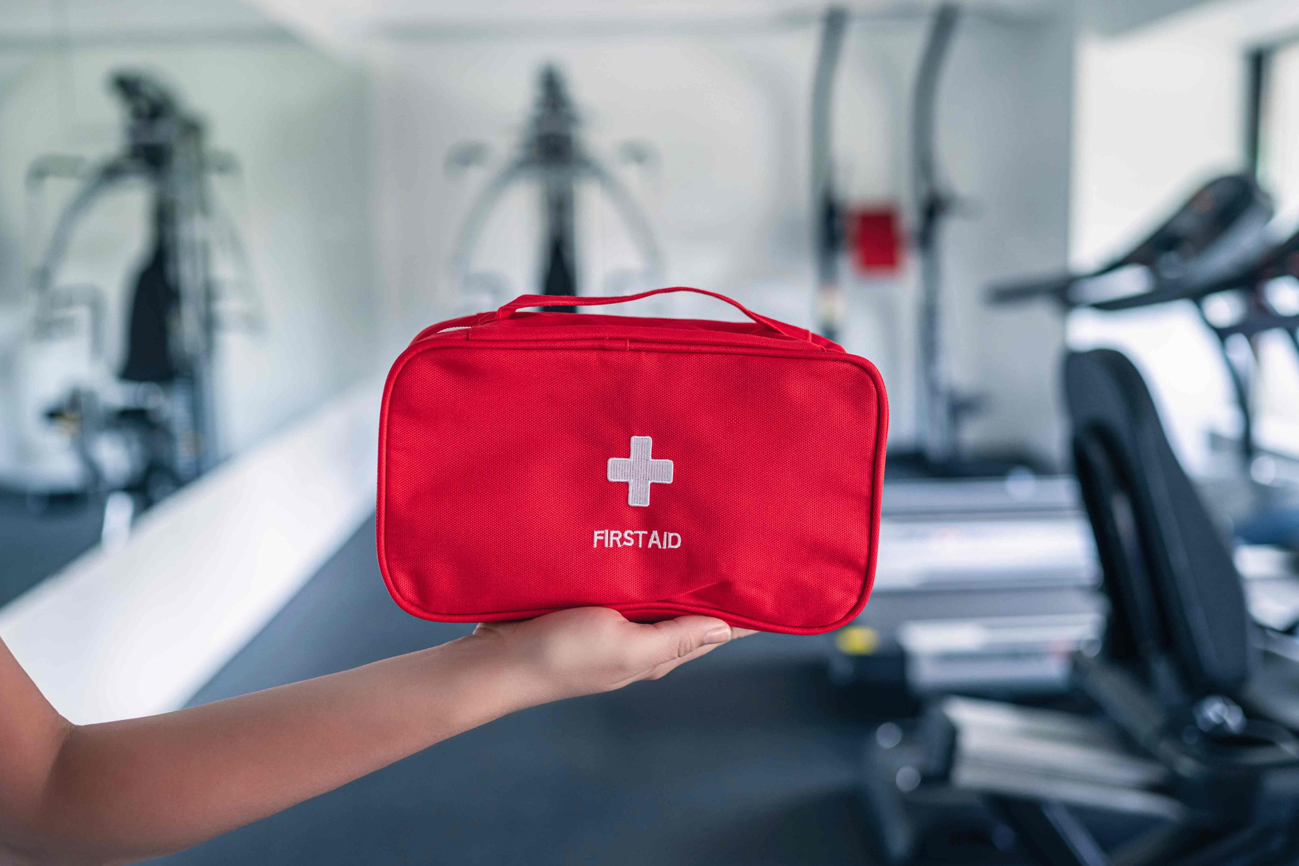 Hand holding a red first aid kit bag with white cross and "FIRST AID" text in a blurred gym setting.